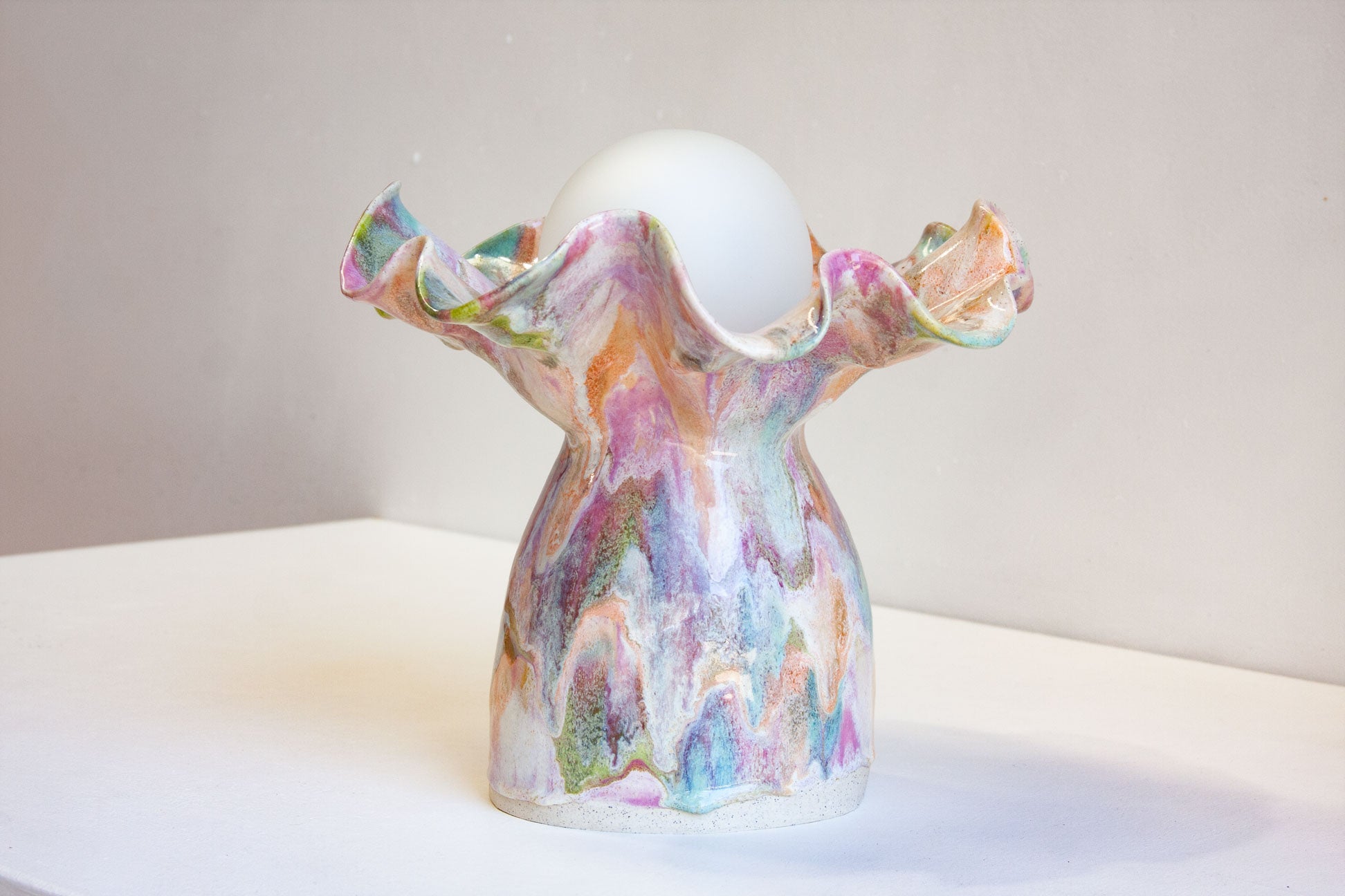 A handmade ceramic candle holder with a ruffled rim and a marbled, multicolored pattern of pink, purple, blue, and green on a white base, holding a white candle on a white surface against a neutral beige background.