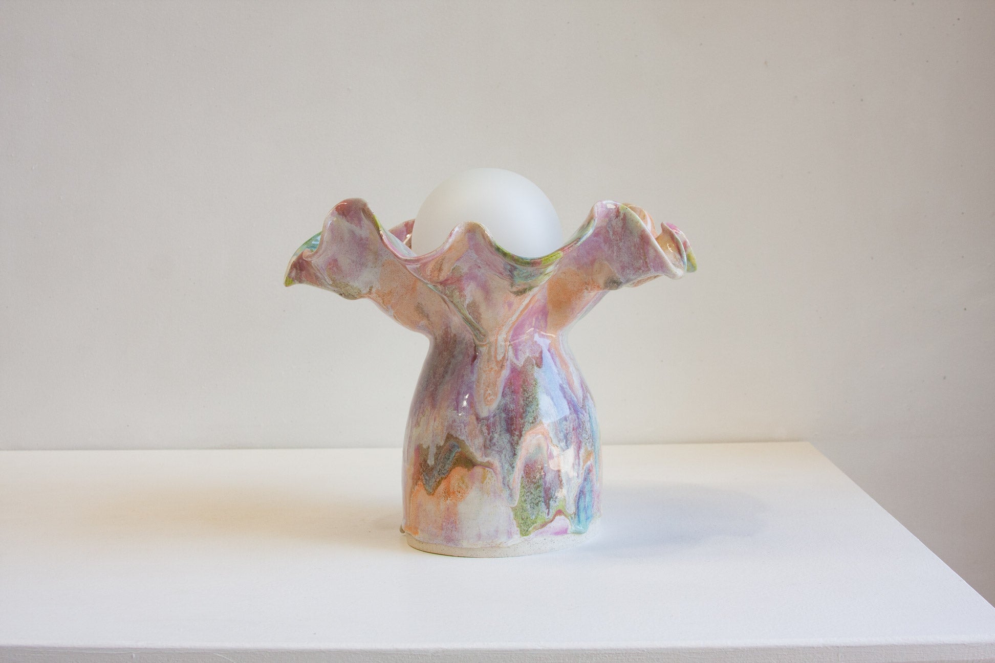 A sculptural ceramic table lamp holder with a flared, ruffled top and a bulb-shaped base, decorated in a marbled pastel glaze of pink, purple, green, and gold, holding a white globe bulb, set against a plain off-white background.