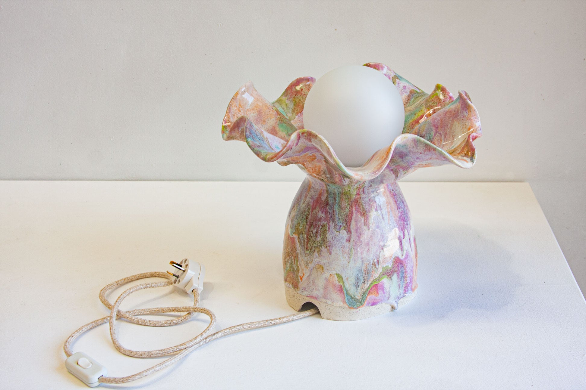 A handmade ceramic table lamp with a ruffled, flower-like rim and a glossy marbled body in pink, green, and cream, featuring a white globe bulb and a coiled white cord with a UK plug on a white surface.