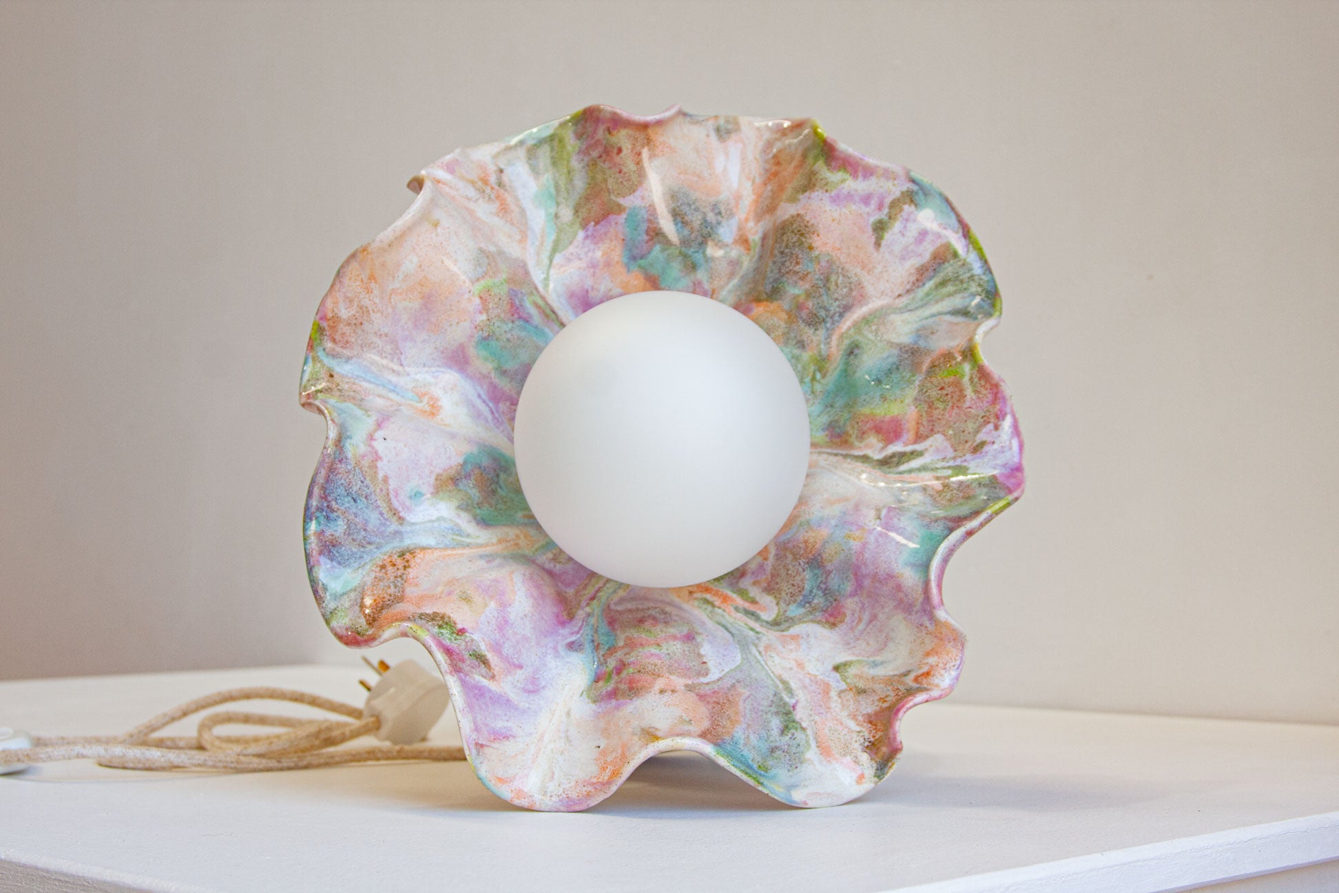 A decorative table lamp with a scalloped, flower-like shade in a marbled pastel pattern of pink, green, blue, and gold, featuring a central white globe bulb. A coiled cord with a plug is visible on the left, resting on a white surface against a neutral beige background.