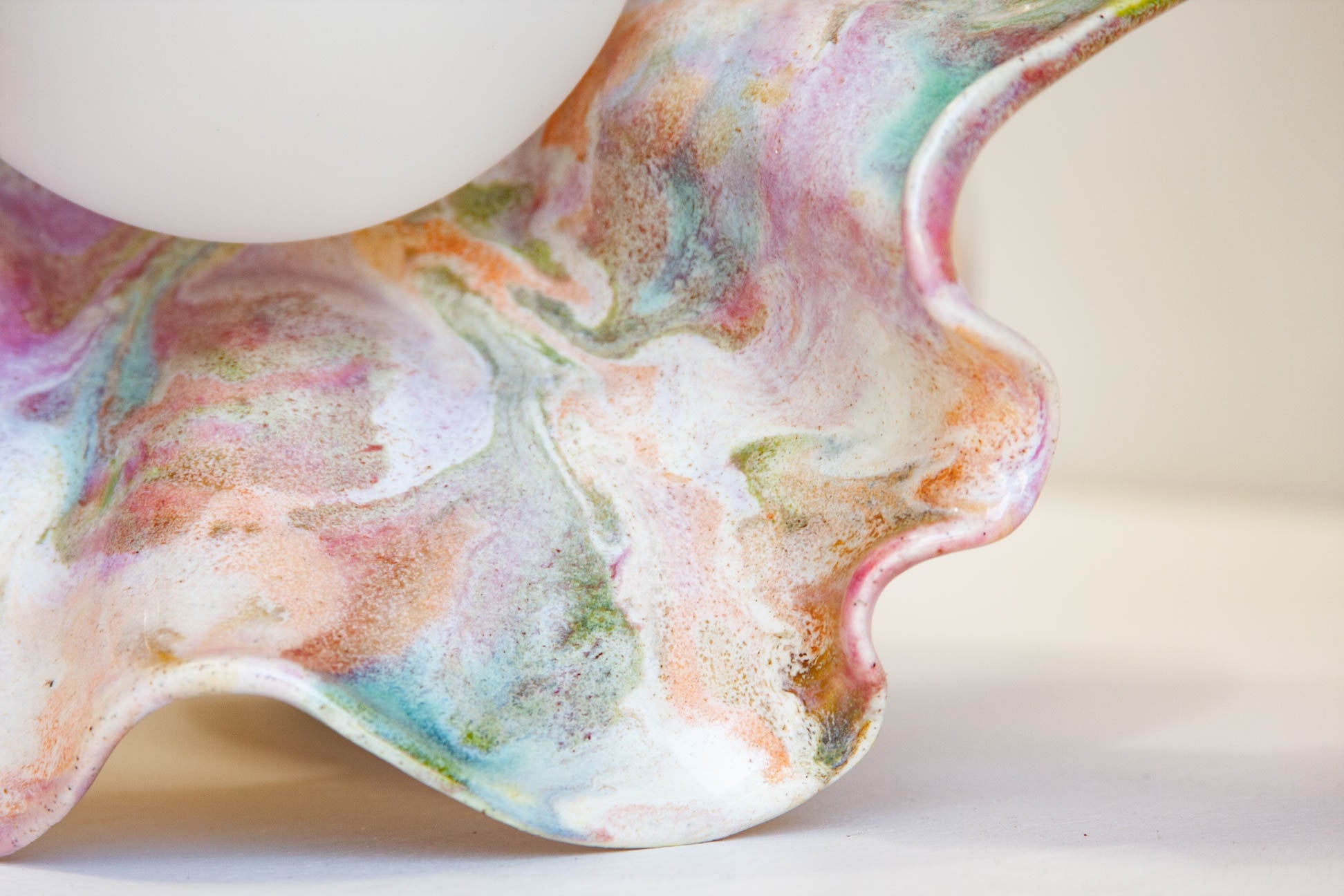 A close-up of a sculptural, organic-shaped ceramic vessel with a marbled, fluid-art finish in pastel pink, purple, green, blue, and orange on a white base, with a white spherical object partially visible at the top.