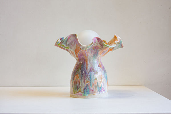 A sculptural ceramic vase with a glossy, marbled multicolor glaze in shades of pink, green, blue, and orange, featuring a flared rim and a white spherical insert at the top, displayed on a white surface against a neutral off-white background.