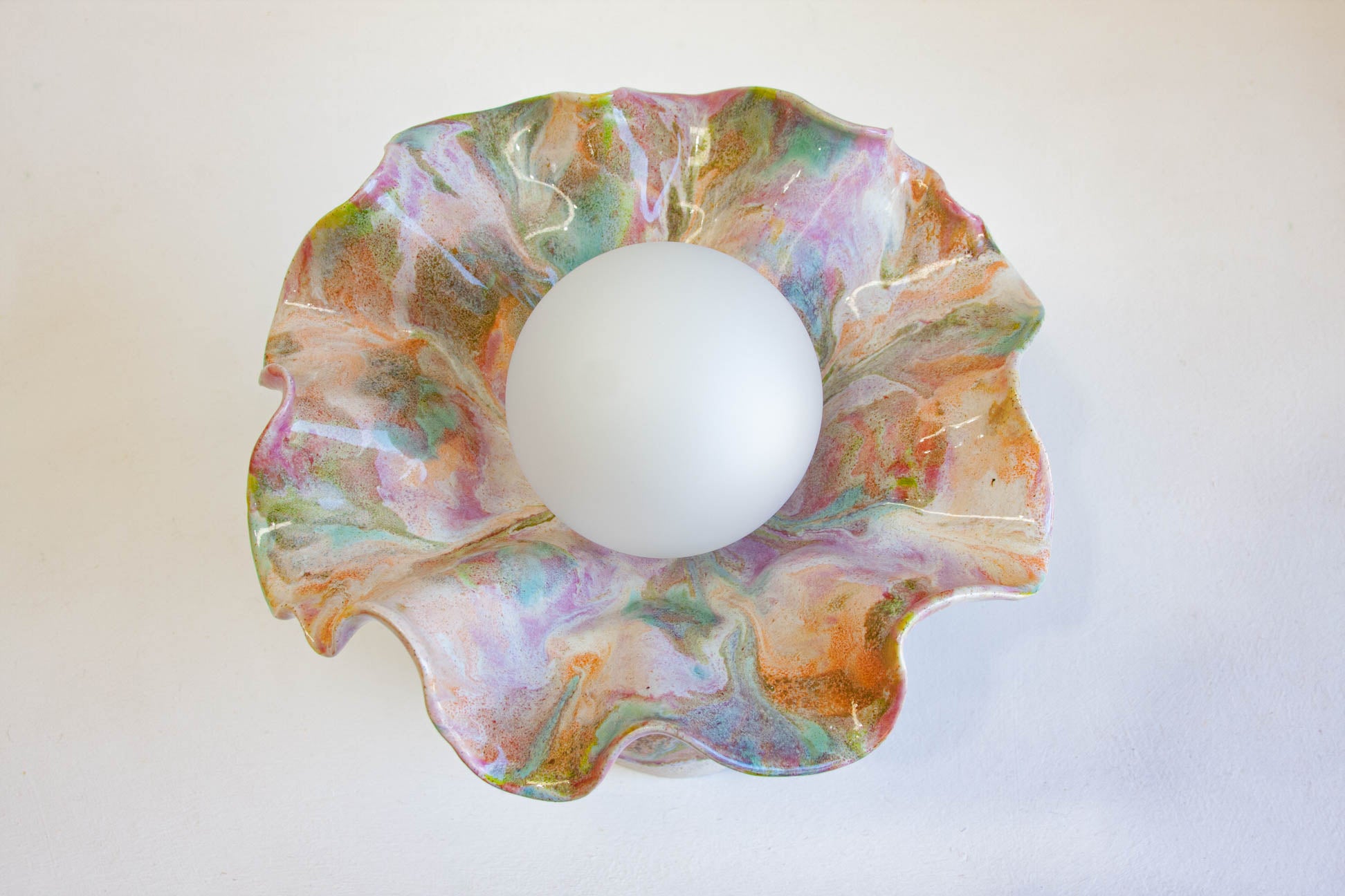 A marbled glass bowl with a scalloped ruffled edge, featuring swirls of pink, green, orange, and white, containing a white frosted glass sphere in the center, set against a plain off-white background.
