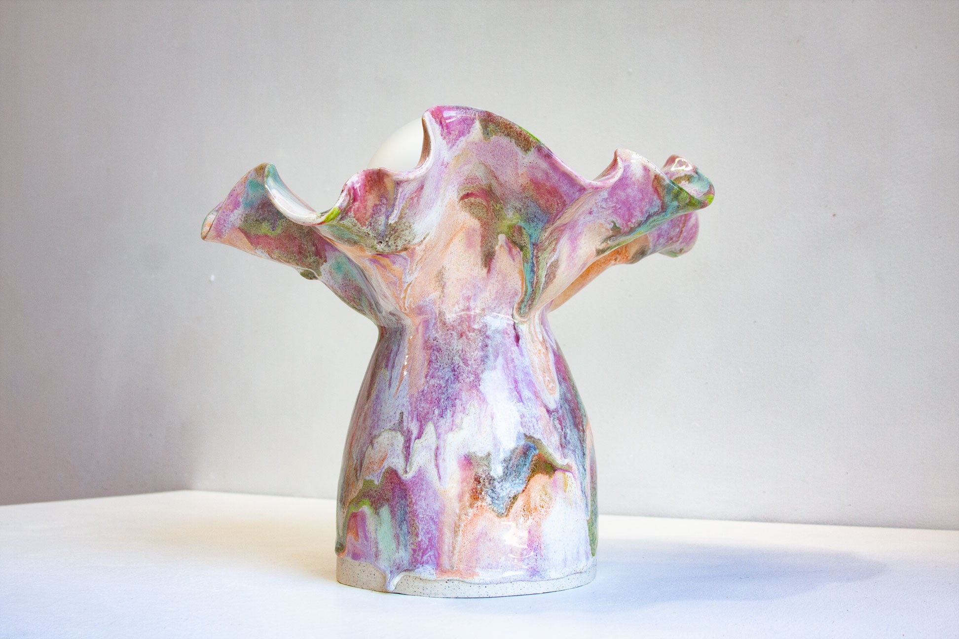 A sculptural ceramic vase with a flared, organic top and a bulbous base, decorated in a marbled glaze of pink, purple, white, green, and brown on a light gray background.