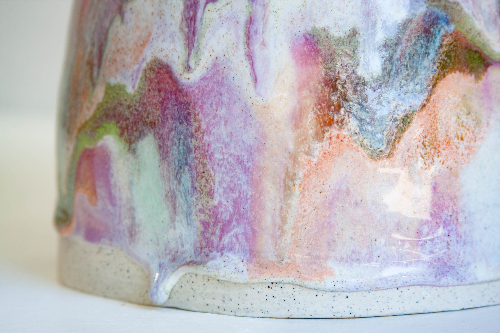 Close-up of a ceramic vessel showing a multicolored dripping glaze with pink, purple, peach, blue, and green tones over a speckled off-white base.