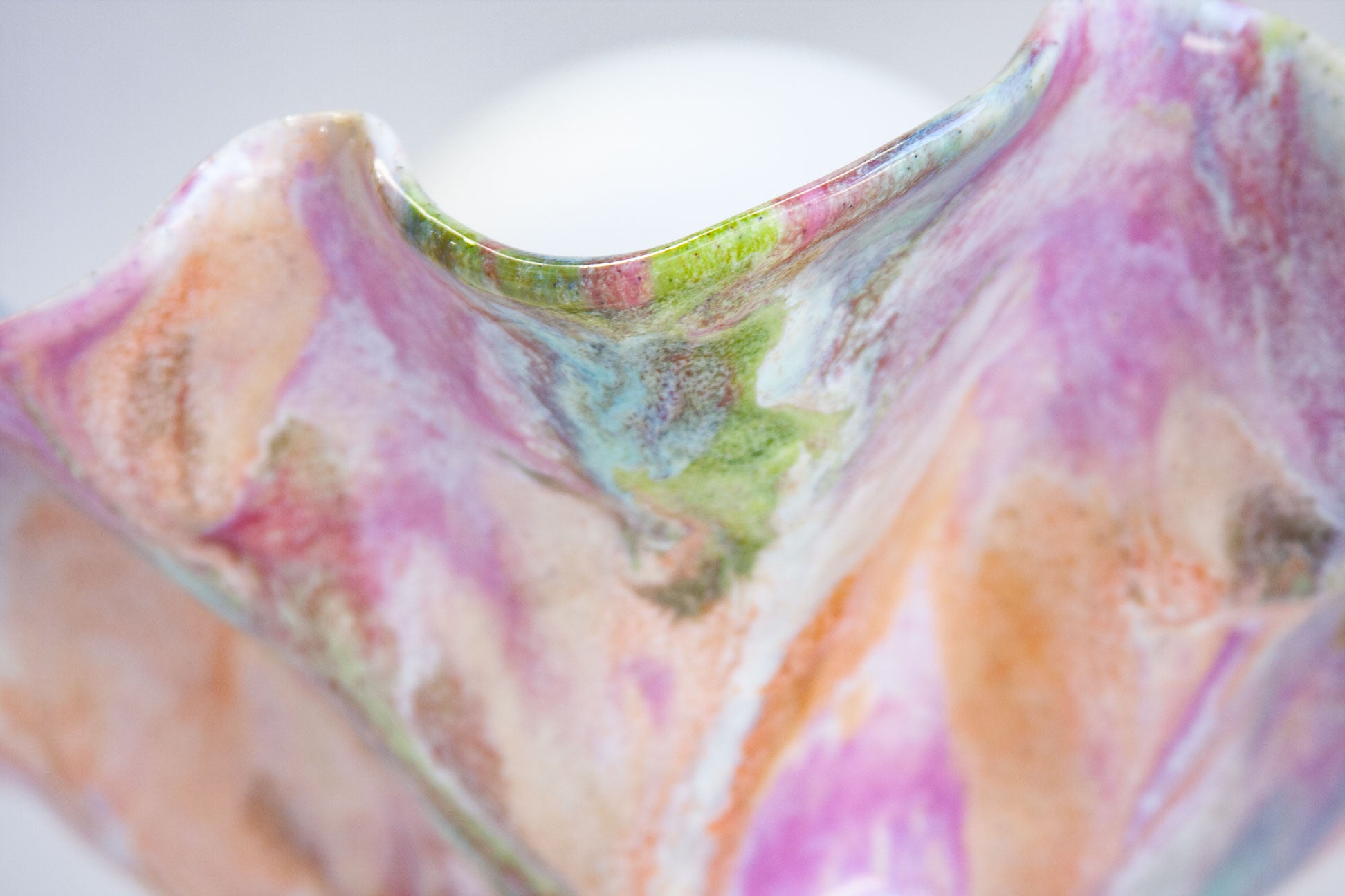 A close-up of a handcrafted ceramic vessel with a wavy rim, decorated in a marbled pattern of pink, purple, green, and orange glazes on a white base.