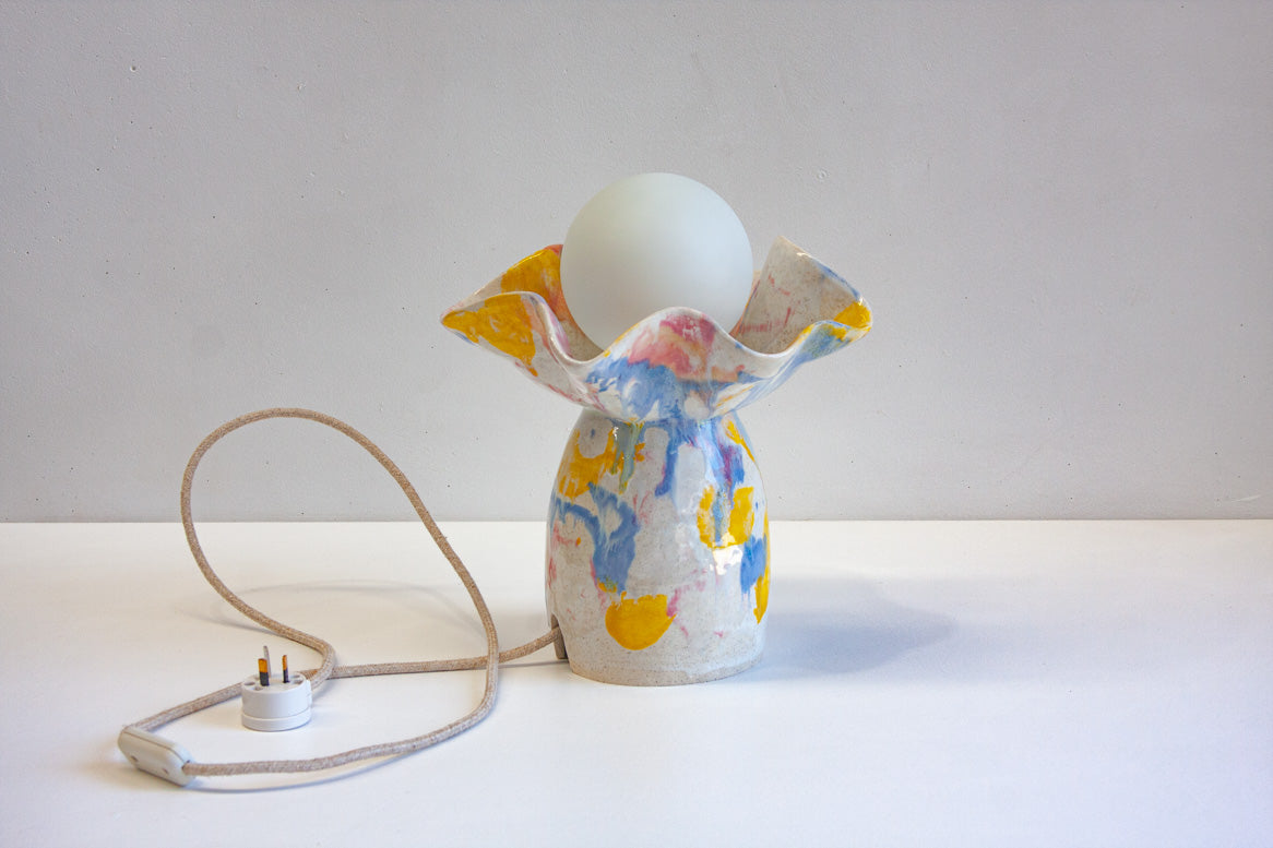 Front-on full-product view of handmade ceramic oyster lamp on a white surface, round-bottomed base with ruffled petal rim glazed in bold yellow, blue, pink and orange on white, white globe bulb, fabric cord with plug laid in front