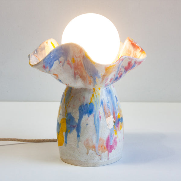 Front-on full-product view of handmade ceramic oyster lamp on a white surface, round-bottomed base with ruffled petal rim glazed in bold yellow, blue, pink and orange on white, white globe bulb, fabric cord with plug laid in front