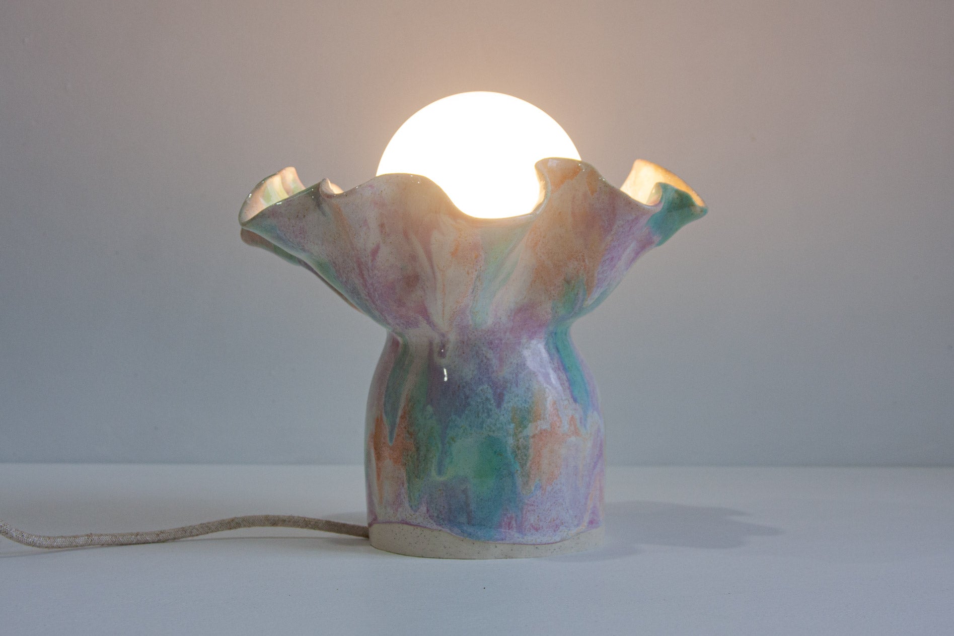 A sculptural ceramic table lamp with a glowing white globe bulb inside a flared, pastel marbled base on a light gray background.