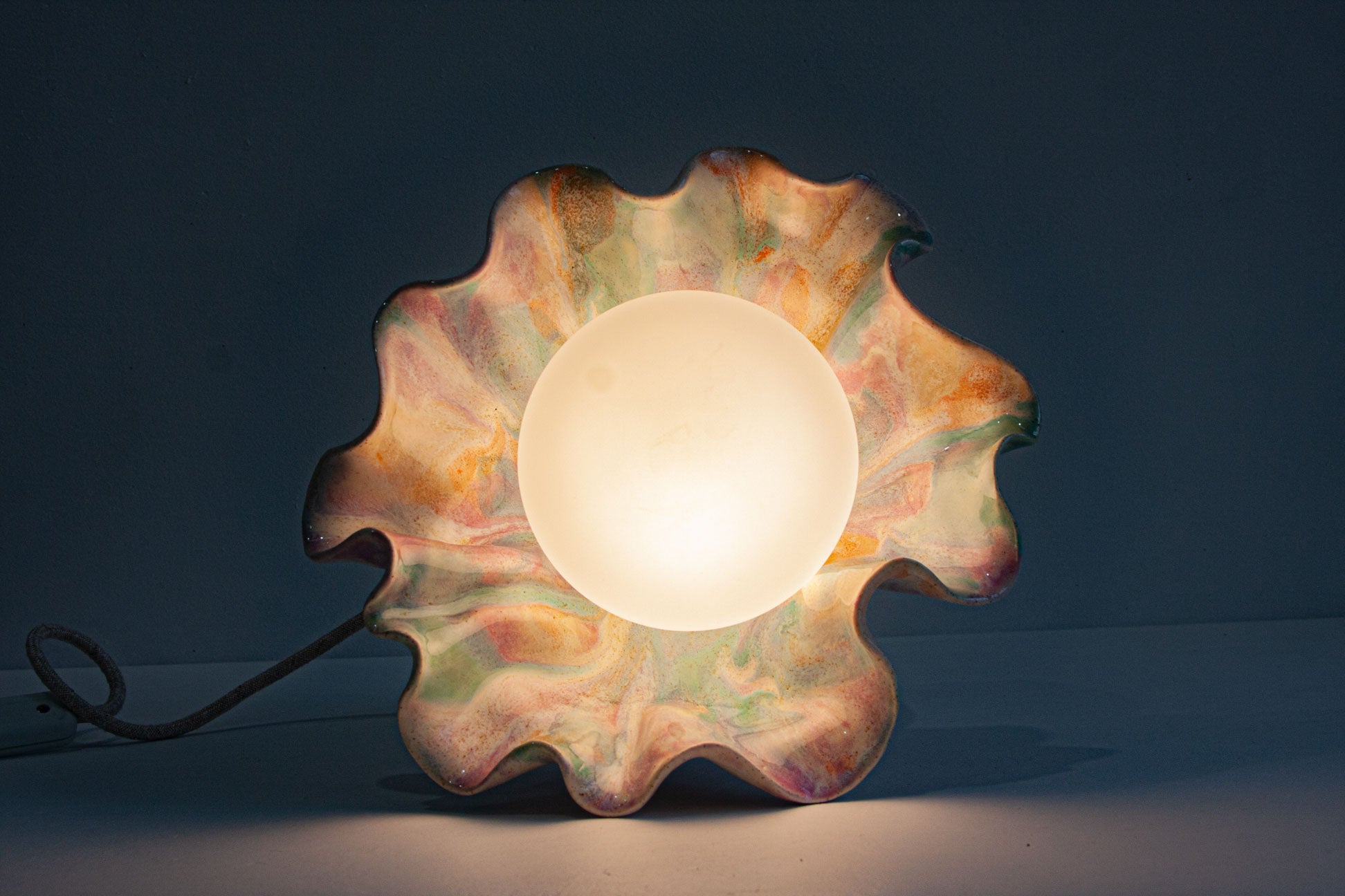 A decorative table lamp with a glowing frosted oval center and a scalloped, marbled outer shade in warm peach, orange, cream, and muted green tones, shown against a dark blue background.