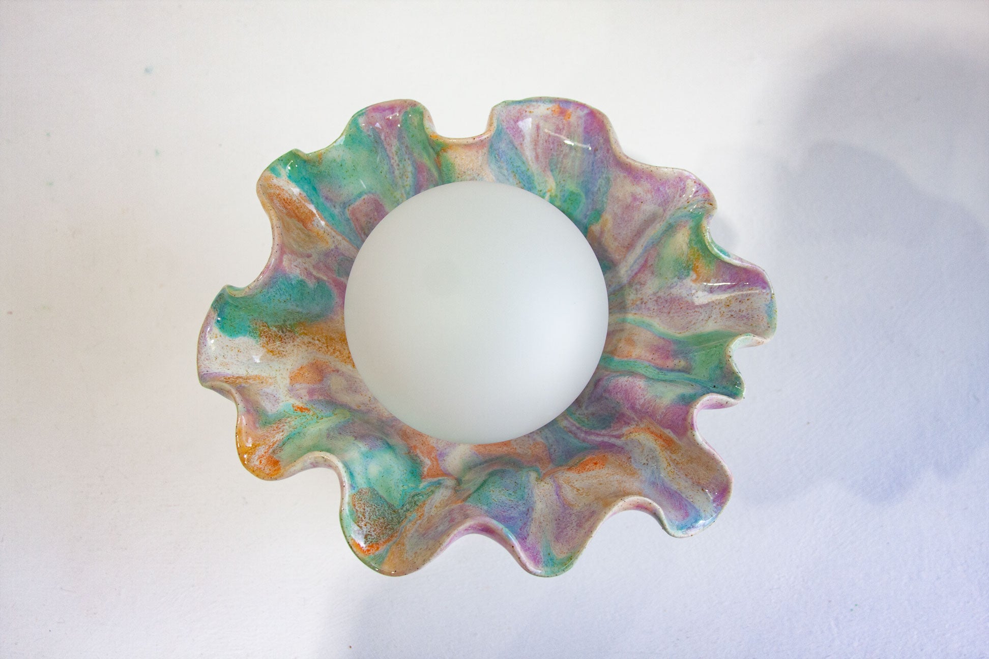 A scalloped, wavy ceramic dish with a marbled pattern of green, purple, pink, and orange, containing a smooth white sphere in the center, set against a plain white background.