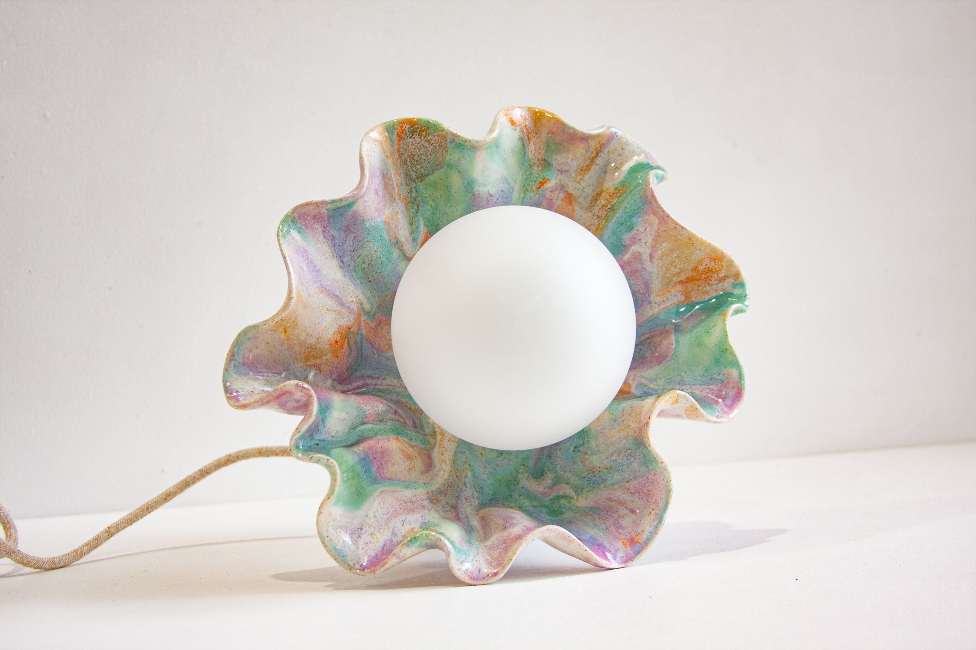 A flower-shaped decorative lamp with a wavy, scalloped rim in a marbled iridescent finish of green, purple, gold, and pink, featuring a central white globe bulb and a visible jute cord on a white background.