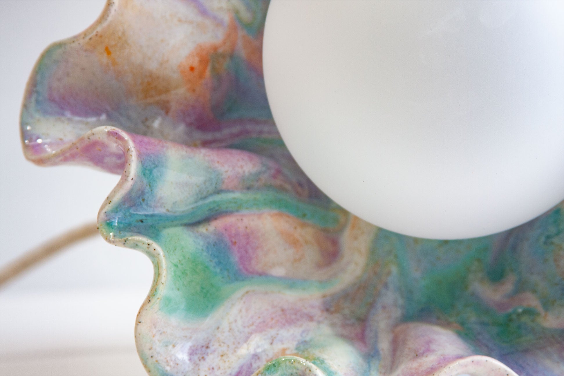 Close-up of a decorative lamp shade made from a clam shell with a marbled, iridescent glaze in pastel purple, green, blue, and orange tones, with a white globe bulb partially visible.