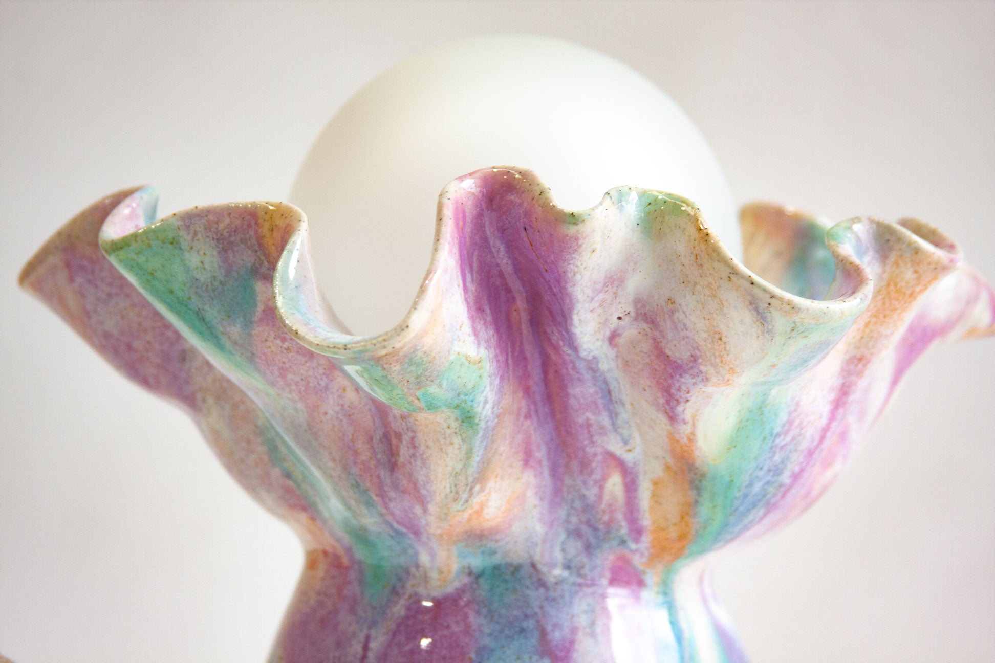 A close-up of a sculptural ceramic vase or candle holder with a flared, ruffled rim, featuring a marbled, iridescent glaze in pastel purple, green, blue, and peach tones. A white spherical object is partially visible behind the top opening against a soft light gray background.
