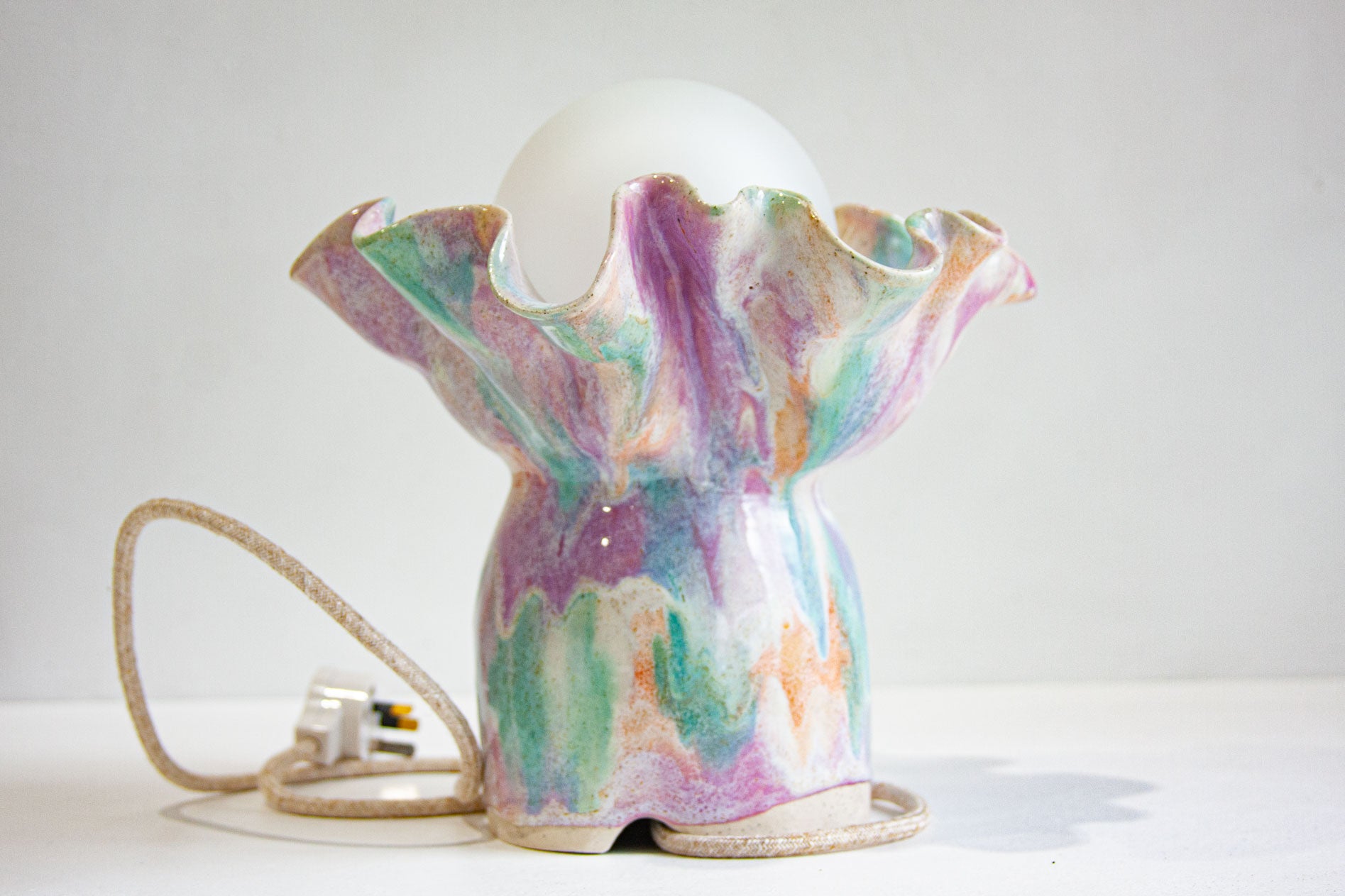 A handmade ceramic table lamp with a ruffled, flower-like base in a marbled pastel glaze of purple, green, pink, and gold, holding a white frosted globe bulb. A coiled beige cord with a UK plug is visible on the left, set against a plain light gray background.