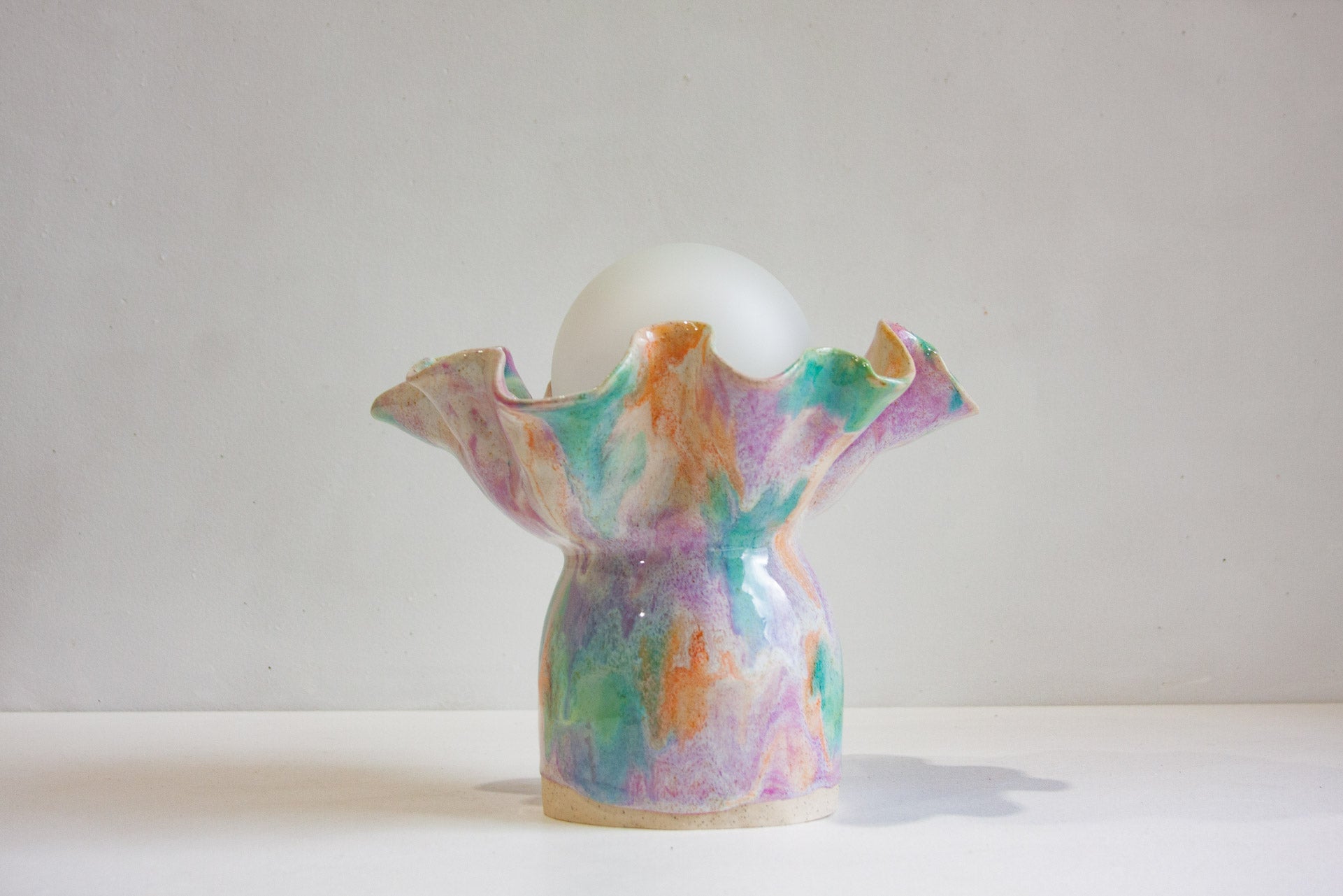 A decorative ceramic lamp holder with a ruffled rim and a glossy marbled pastel finish in purple, green, orange, and blue, holding a white globe light on a plain light gray background.