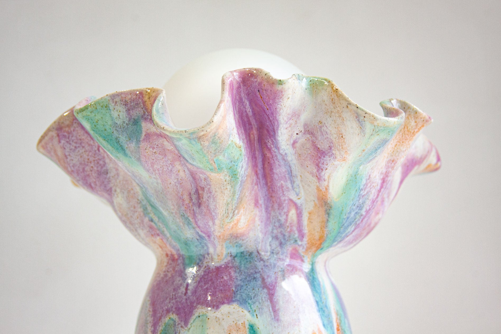 A close-up of a sculptural ceramic vase with a flared, ruffled rim and a glossy marbled finish in pastel purple, mint green, peach, and white, with a white spherical top insert.