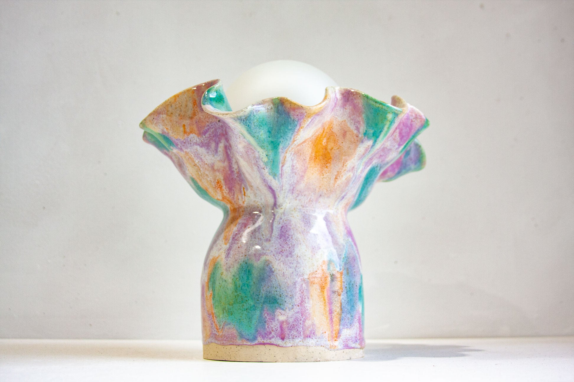 A handmade ceramic table lamp with a sculptural, flared base and a smooth white spherical bulb, decorated with a multicolored marbled glaze of purple, green, orange, and pink on a light background.