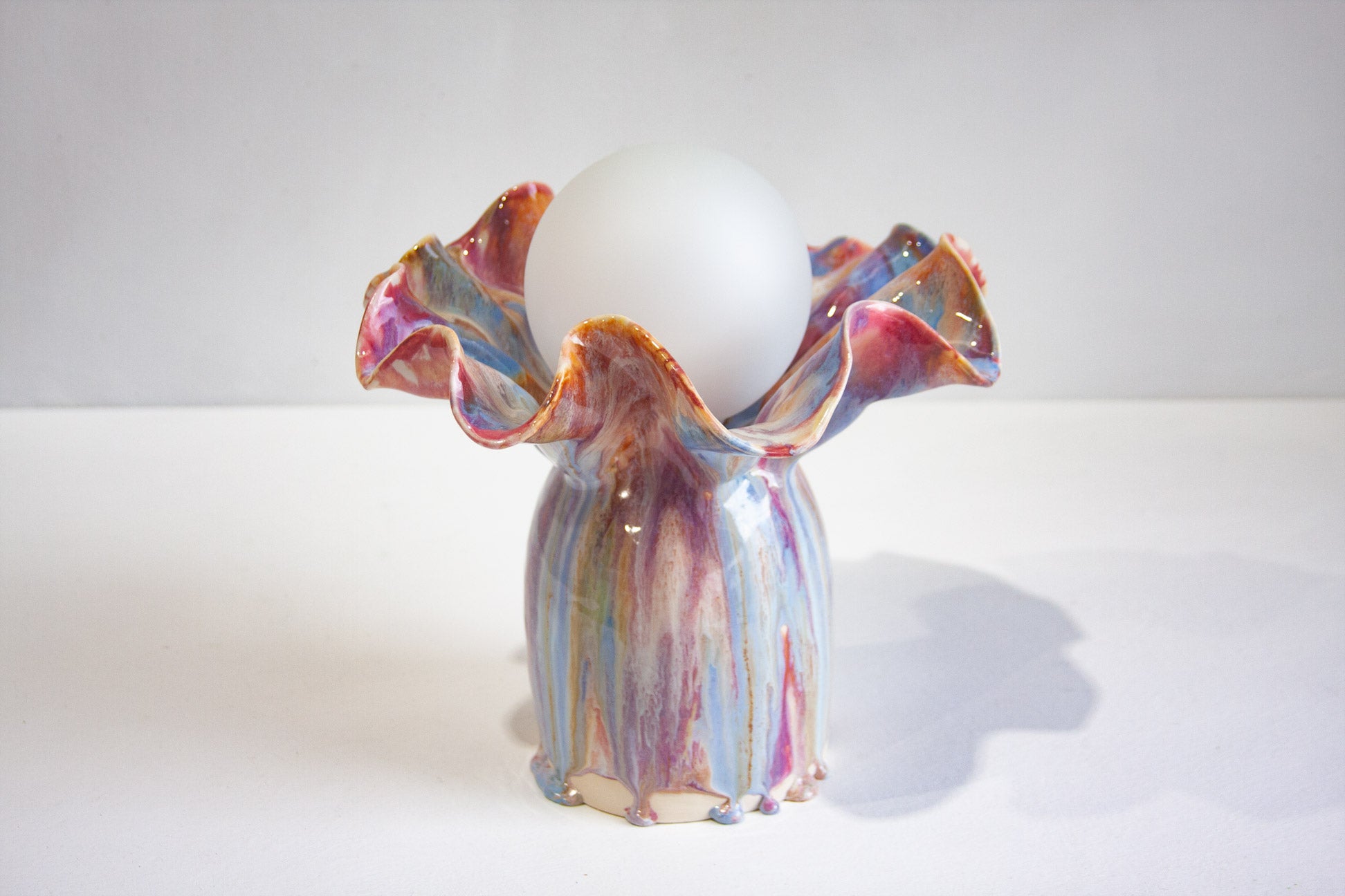 A handmade ceramic lamp with a ruffled, iridescent rim and a matching glazed base, holding a white ceramic globe on a white surface against a light gray background.