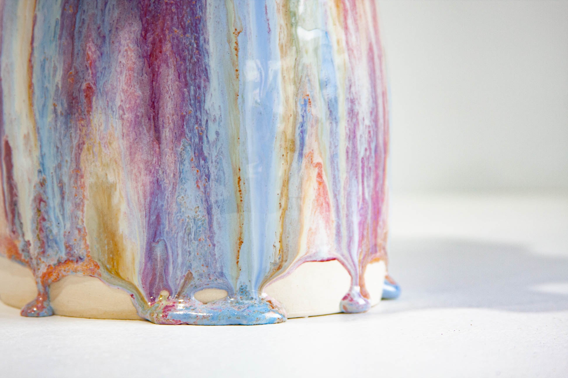 A close-up of a ceramic vase with a dripping glaze design featuring vertical streaks of pastel blue, purple, pink, and gold on a white base.