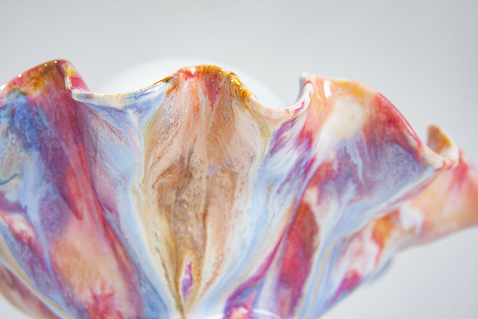 A close-up of a hand-painted ceramic lamp with a ruffled rim, featuring a marbled pattern of blue, pink, purple, and gold glazes on a light grey background.