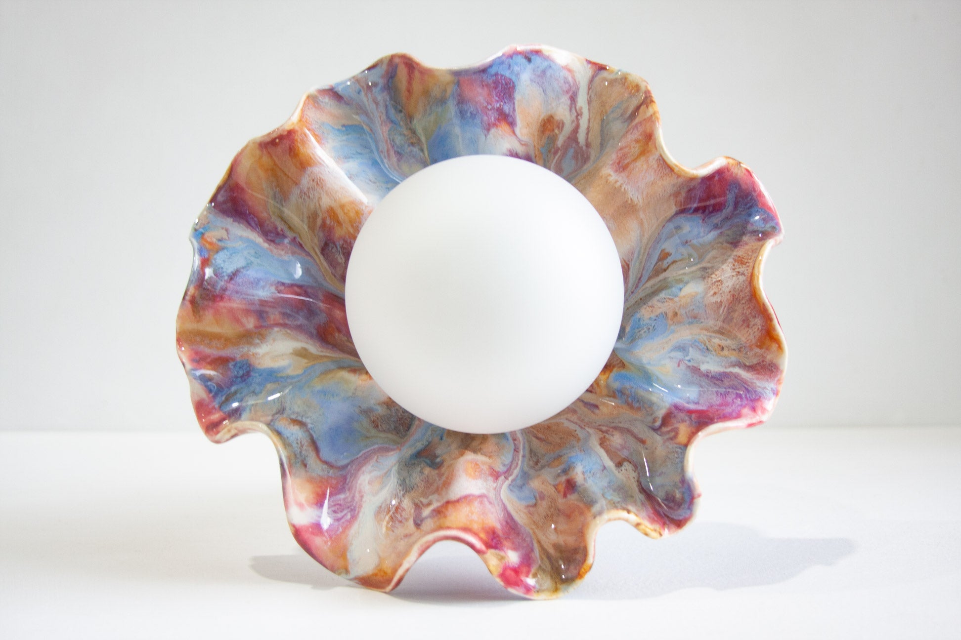 A scalloped ceramic lamp with a marbled pattern of blue, pink, purple, and brown glaze, holding a smooth white bulb in the center, set against a plain white background.