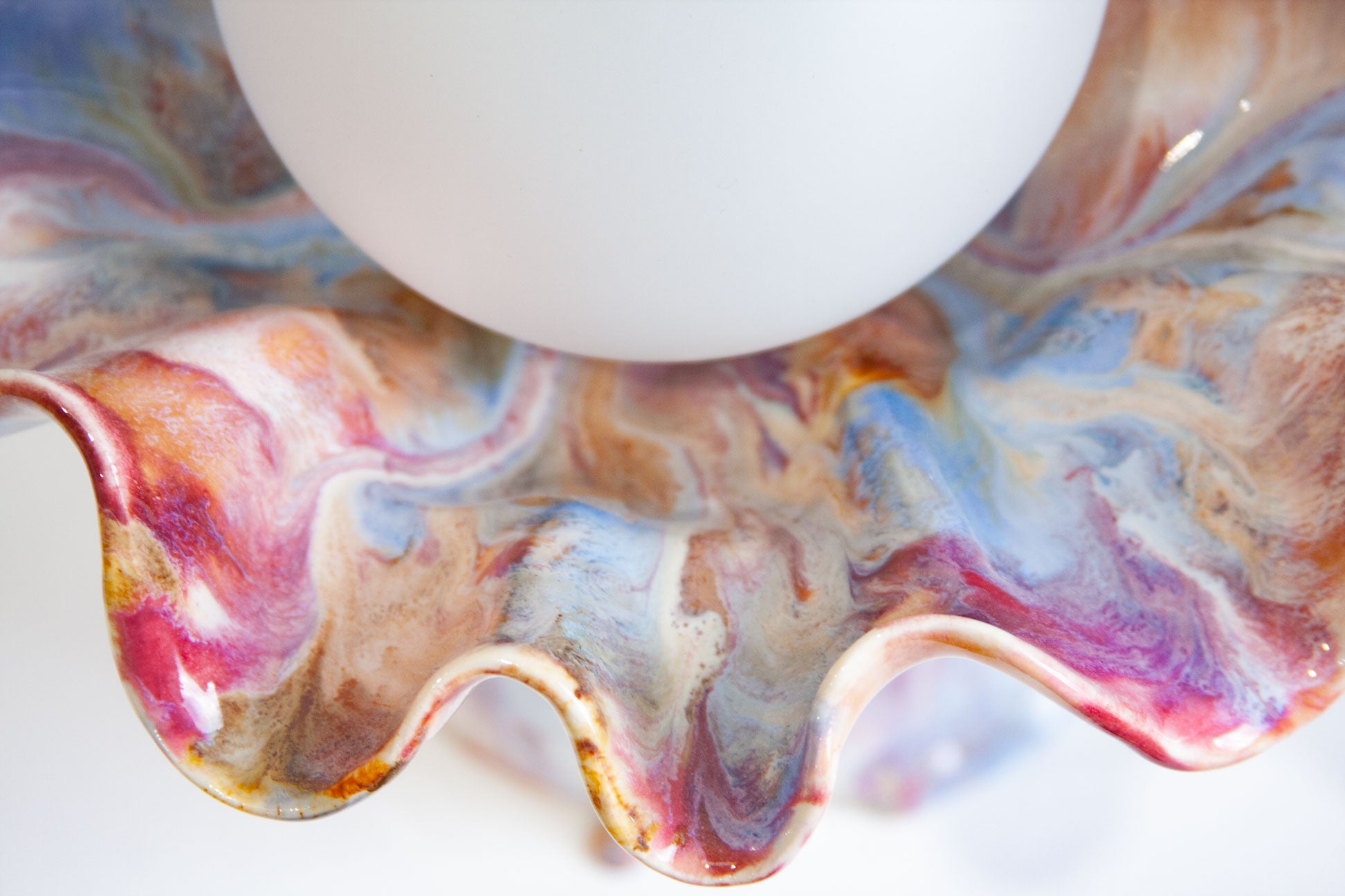 A close-up of a decorative ceramic bowl with a wavy, scalloped rim, featuring a marbled pattern of blue, pink, purple, brown, and white. A white ceramic globe is partially visible at the top.