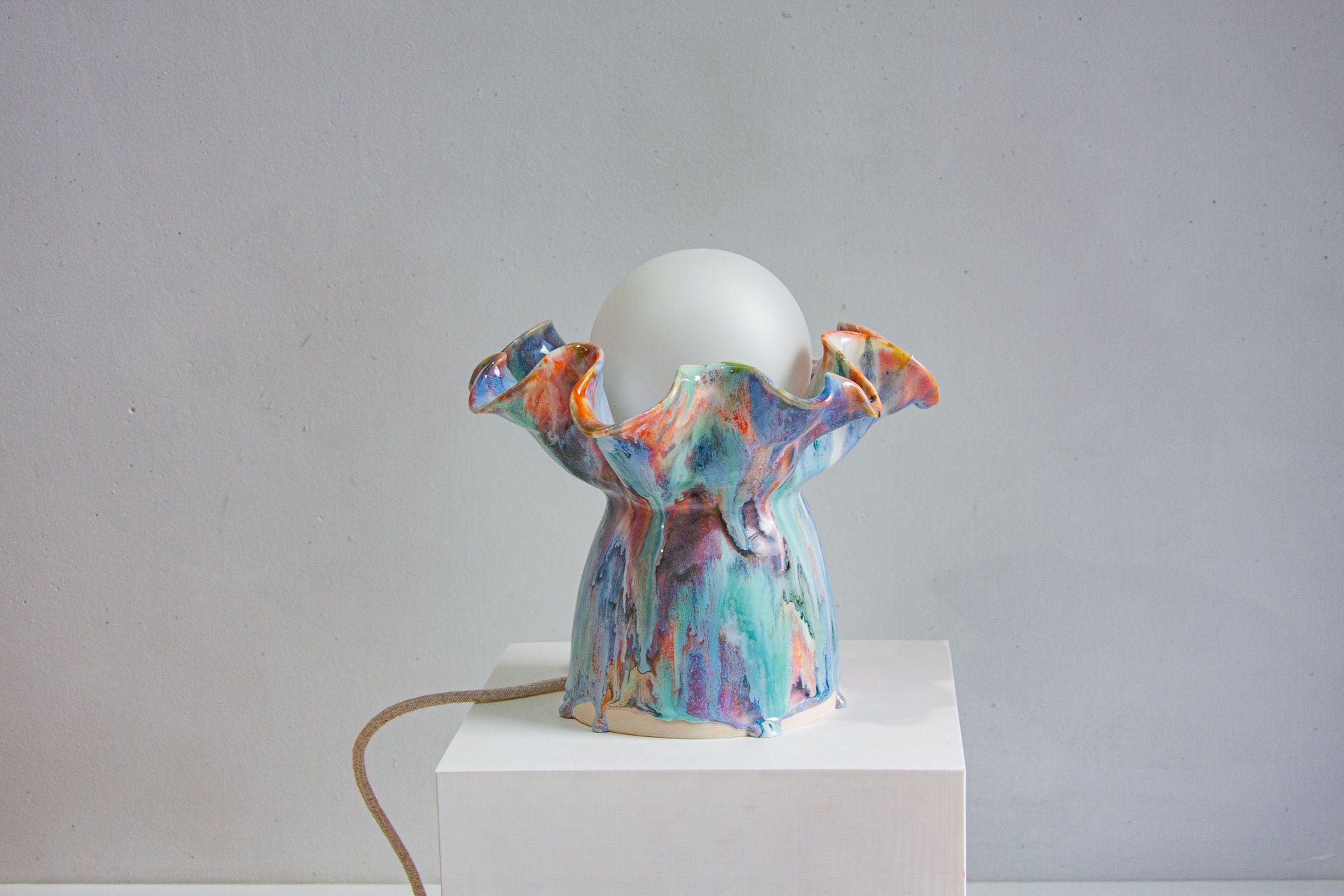 handmade ceramic table lamp with bulb and cable visible, sitting on a white plinth with a white background