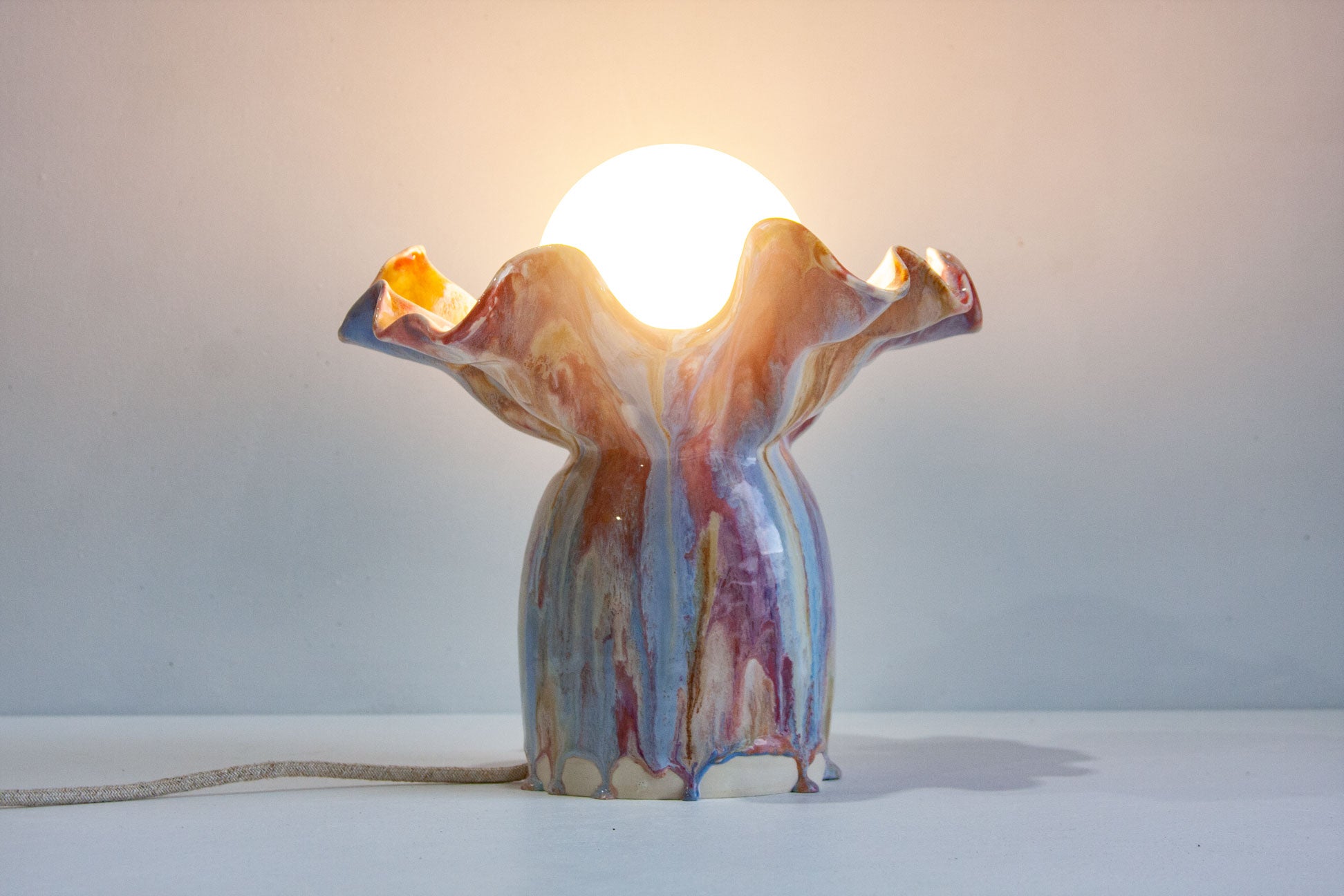 A sculptural ceramic table lamp with a glowing bulb inside, featuring a wavy, flared top and a glossy marbled body in blue, purple, pink, and orange tones, set against a plain light gray background.