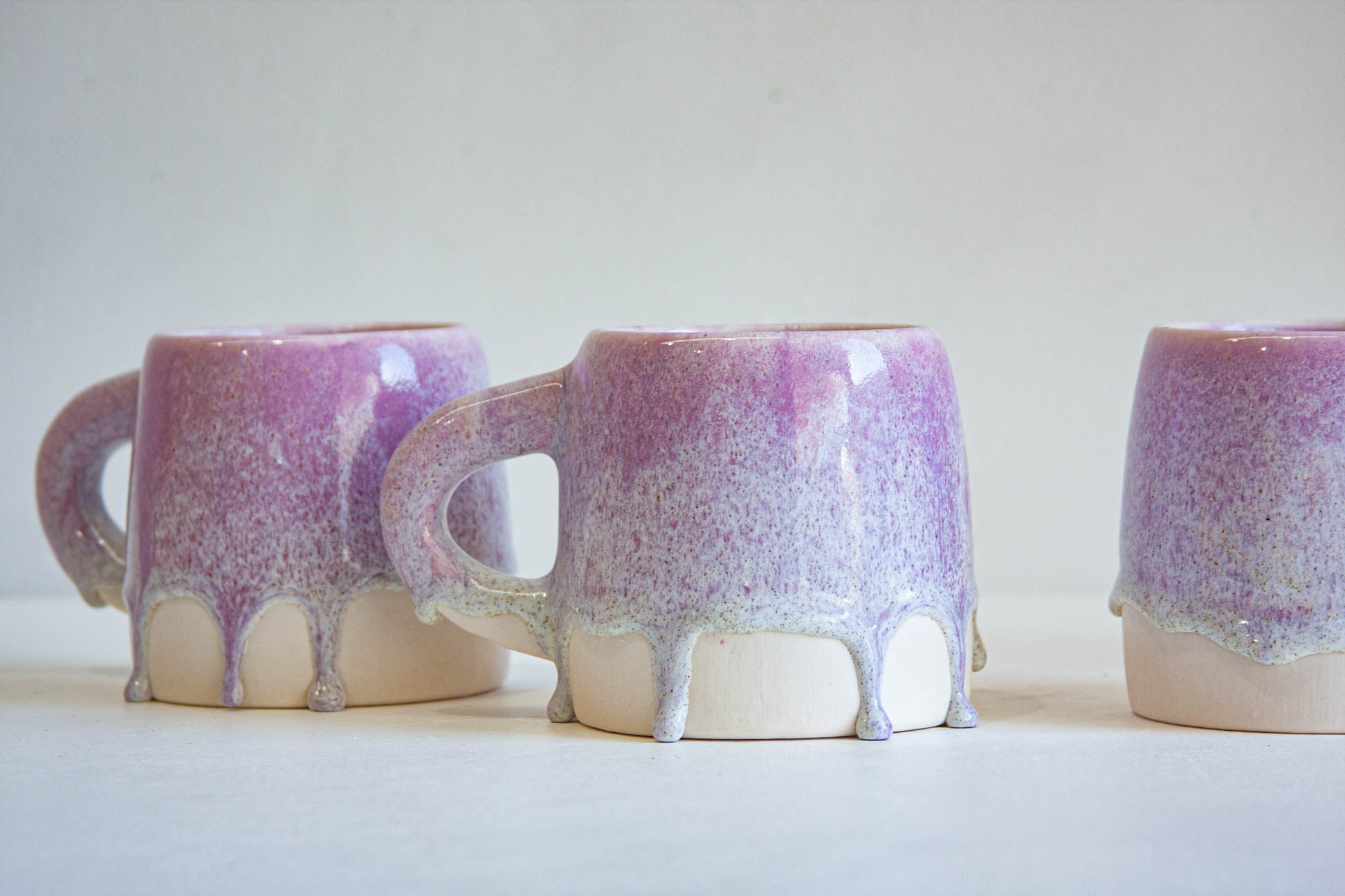 Three ceramic mugs with a purple and lavender glaze dripping over a cream-colored base, arranged on a light surface against a plain light background.