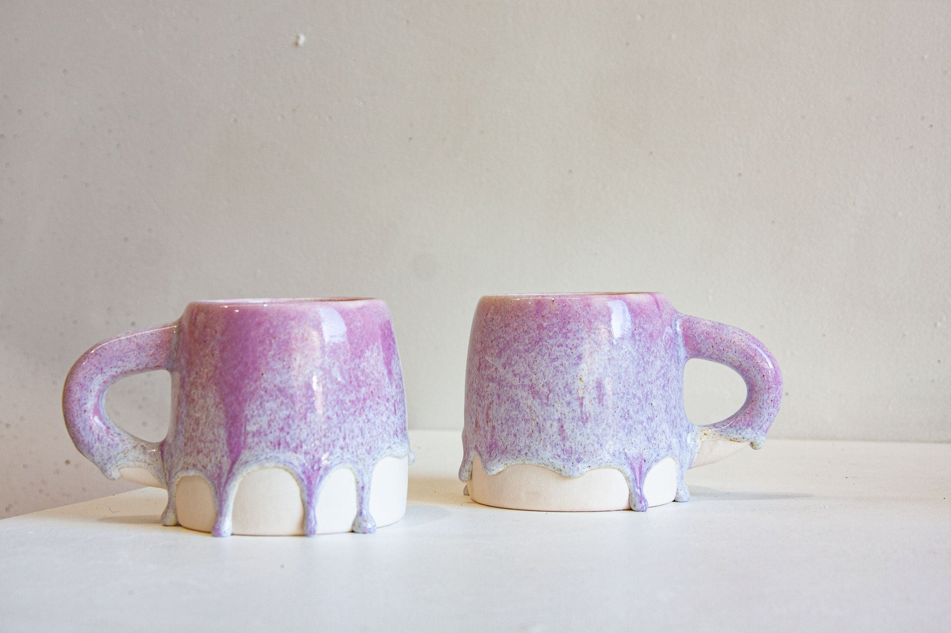 Two ceramic mugs with a purple and lavender drip glaze and white dripped accents at the base, displayed side-by-side on a white surface against a neutral light gray background.