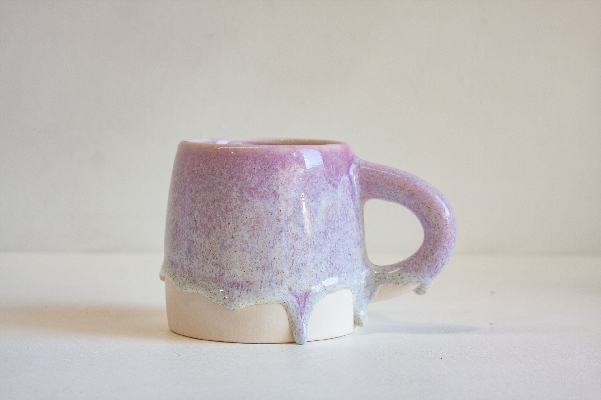 A handmade ceramic mug with a lavender and purple glaze dripping over a white base, set against a plain off-white background.