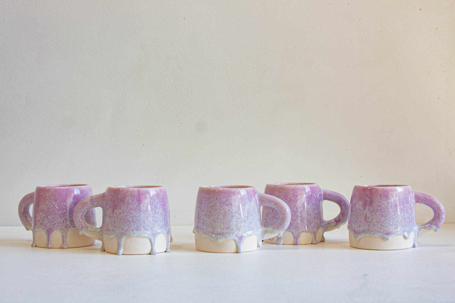Five small ceramic mugs with purple and lavender glaze and dripping white clay at the base, arranged in a row against a plain off-white background.