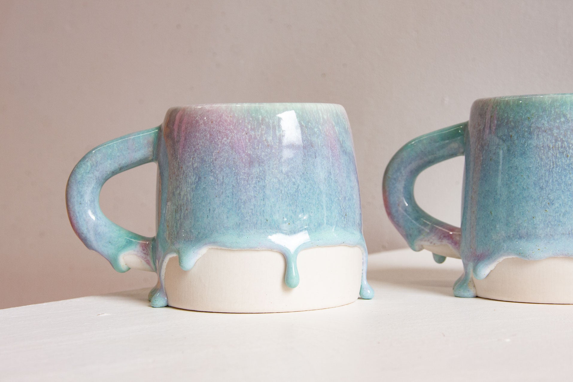 Two handmade ceramic mugs side by side, loop handles facing out, the purple and blue glaze running down to a line of drips over bare white clay.
