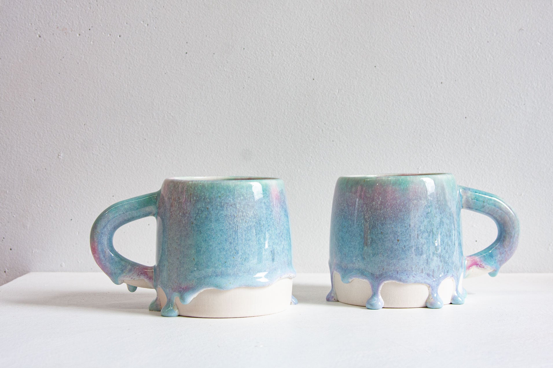 Two handmade ceramic mugs in a light purple and blue glaze, one tipped on its side to show the pale interior, with rounded drips standing in a row along the foot.