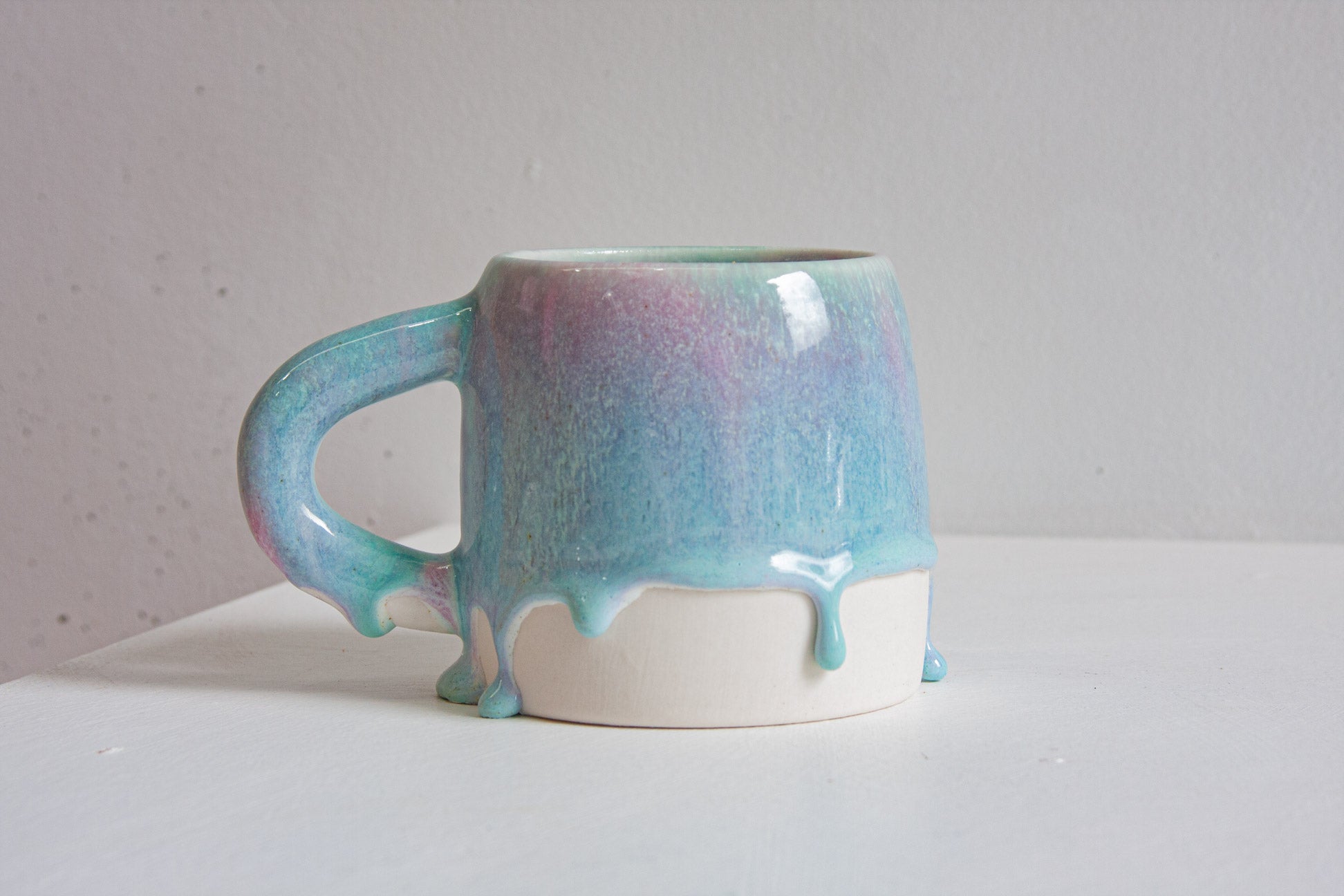 Five handmade ceramic mugs in a row, each in a light purple and blue glaze that drips over the bare white clay base, against a plain wall.