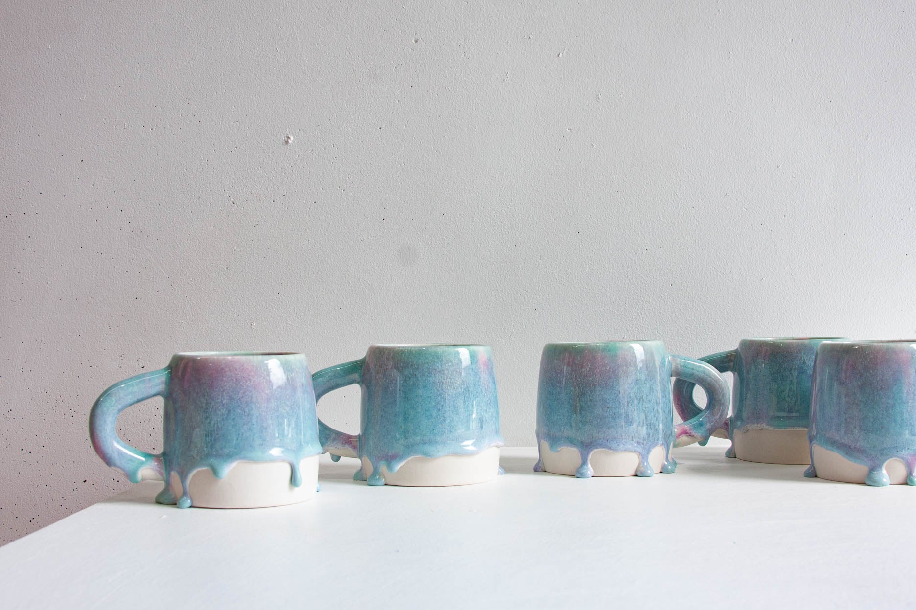 Close-up of two handmade ceramic mugs in a light purple and blue glaze, thinning at the rim and running into rounded drips above the bare white clay base.