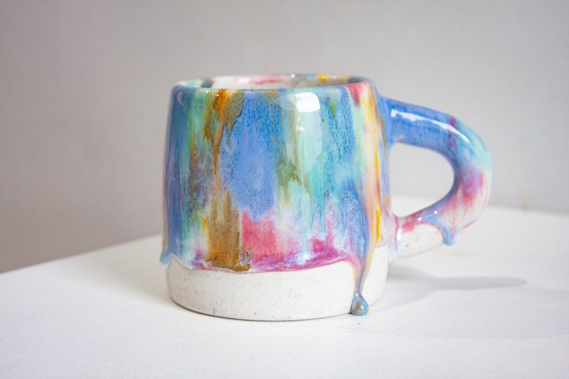 A handmade ceramic large mug seen side on, brush-glazed in pink, yellow, green and blue running down to drips above the bare white clay base.