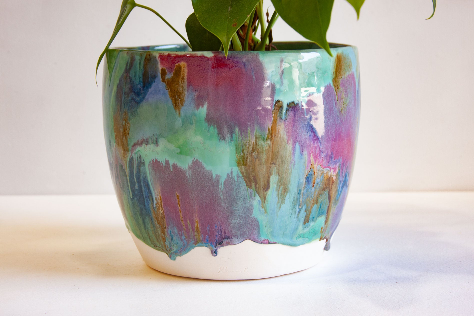 A ceramic plant pot with a colorful, abstract dripping glaze in shades of teal, purple, pink, and gold, containing a green houseplant against a plain white background.