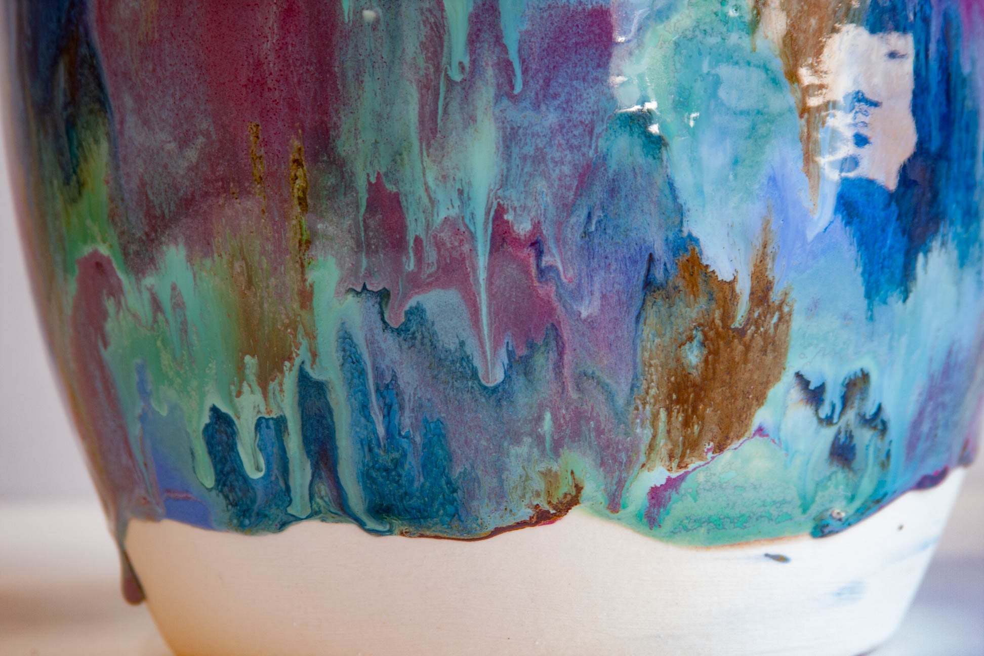 Close-up of a ceramic vase with a glossy drip glaze featuring swirling shades of blue, teal, purple, and brown over a white base.