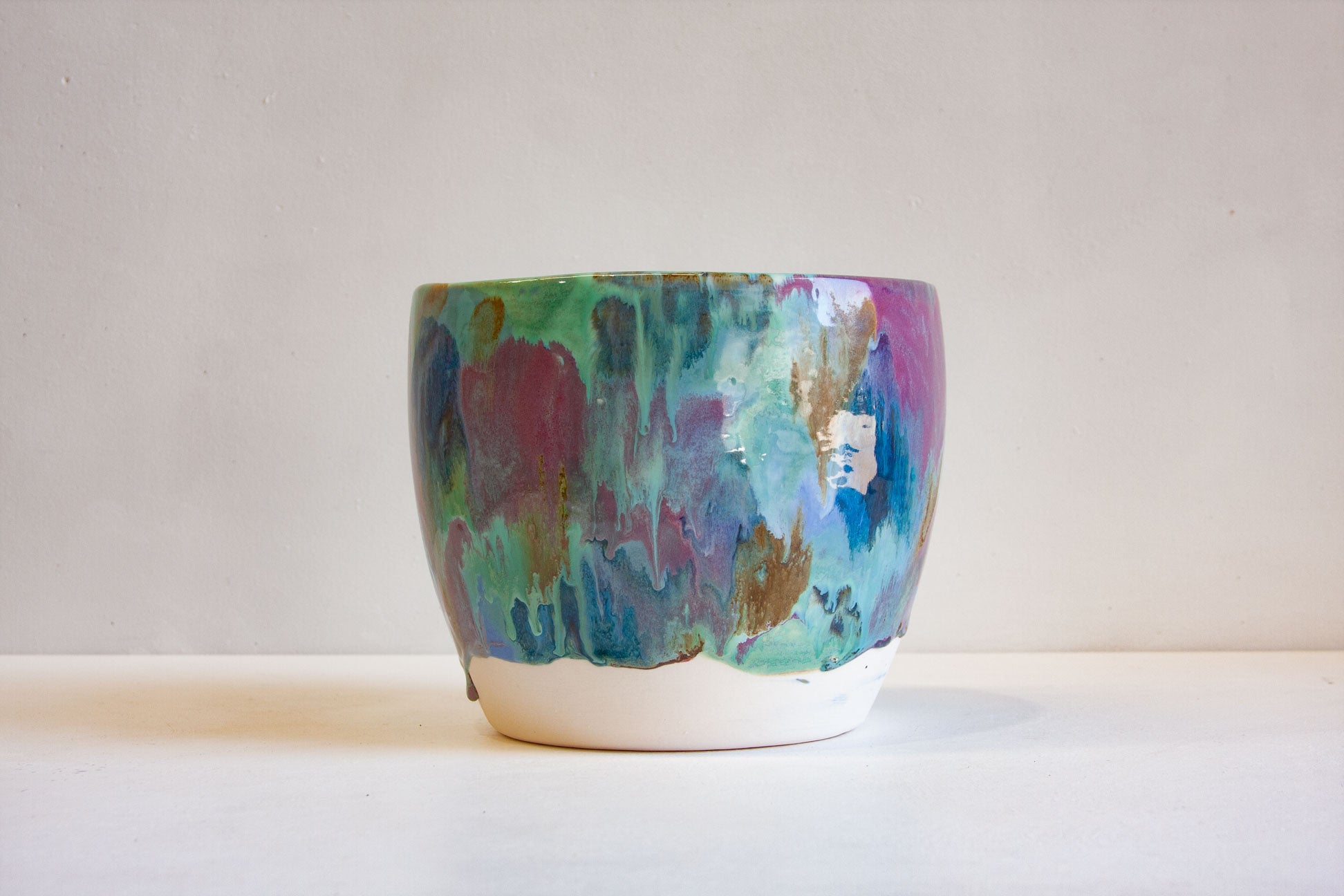 A handmade ceramic planter with an abstract, multicolored painted design in shades of teal, blue, purple, and gold, featuring a raw, white unglazed base, set against a plain off-white background.