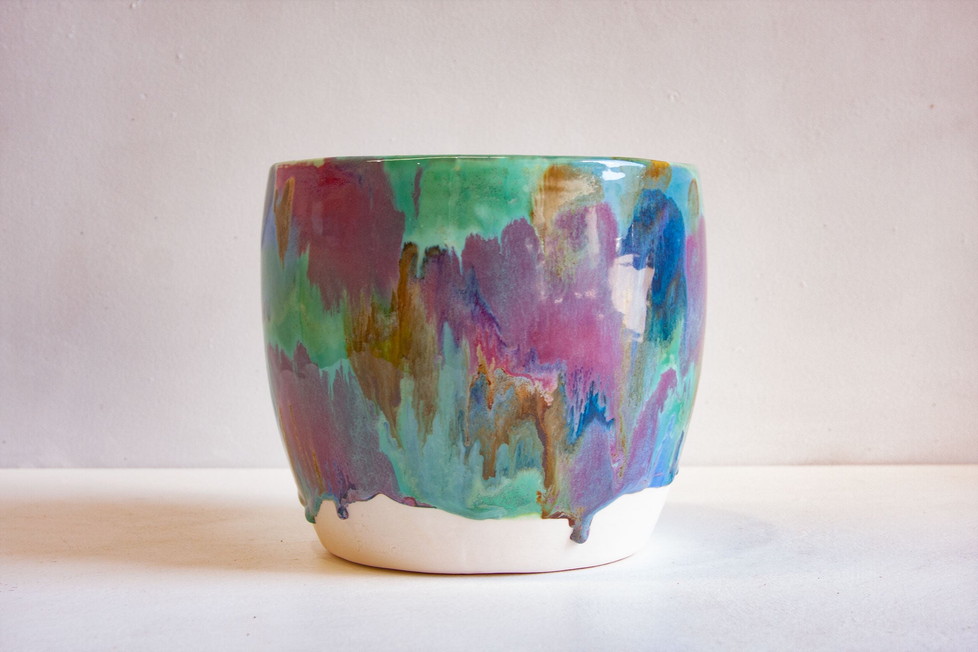 A handmade ceramic planter with a colorful, abstract dripping glaze in shades of teal, purple, pink, and blue, sitting on a white surface against a neutral light gray background.