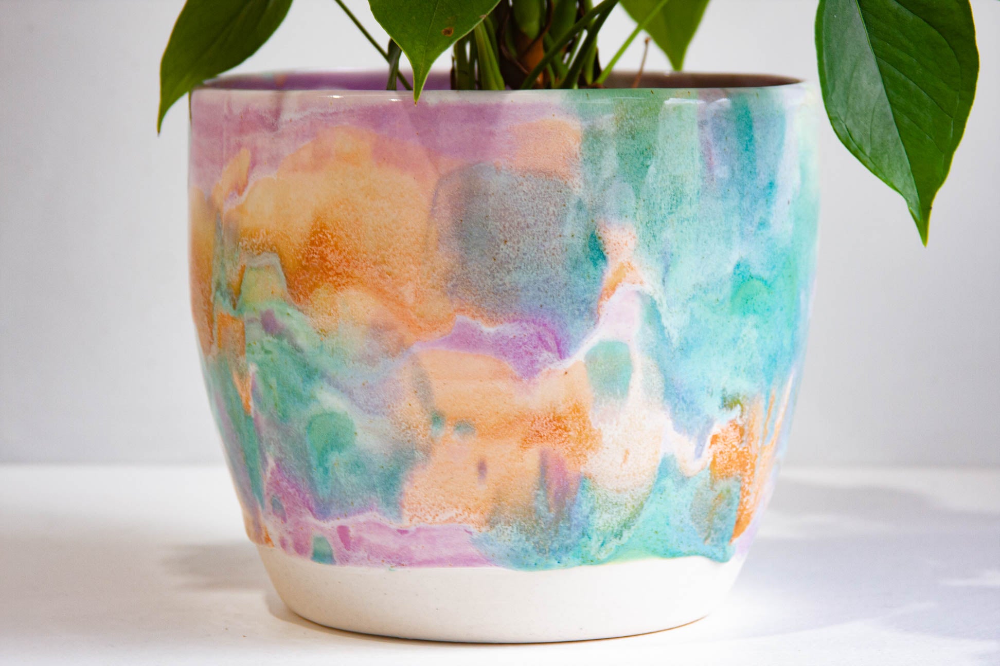 A ceramic planter with a colorful watercolor-style marbled pattern in shades of teal, orange, purple, and pink, with a white glazed base, holding a green plant against a plain white background.
