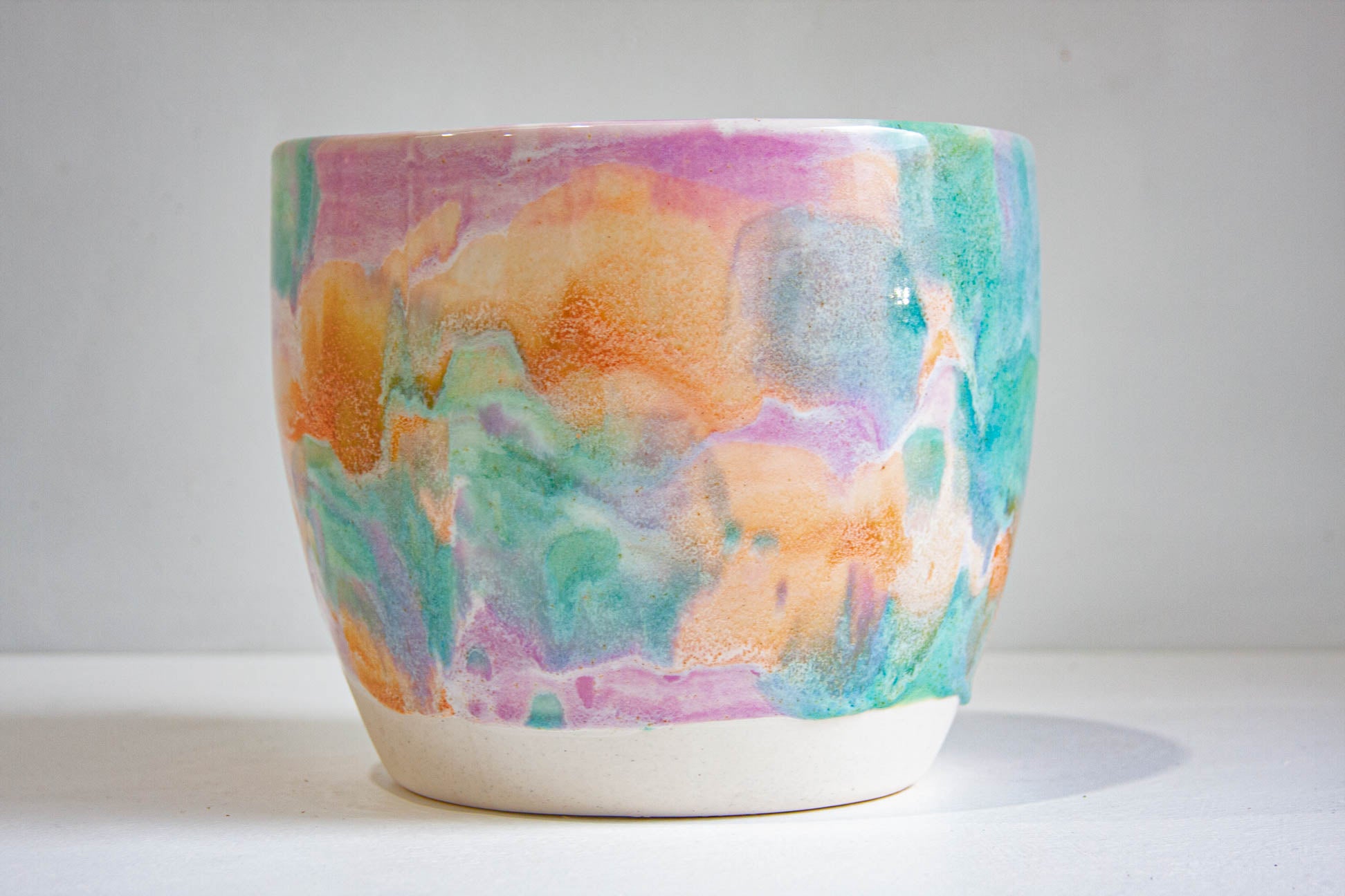 A handmade ceramic planter pot with a colorful, abstract watercolor-like pattern in shades of orange, pink, green, and blue on a white glazed base, set against a plain light gray background.