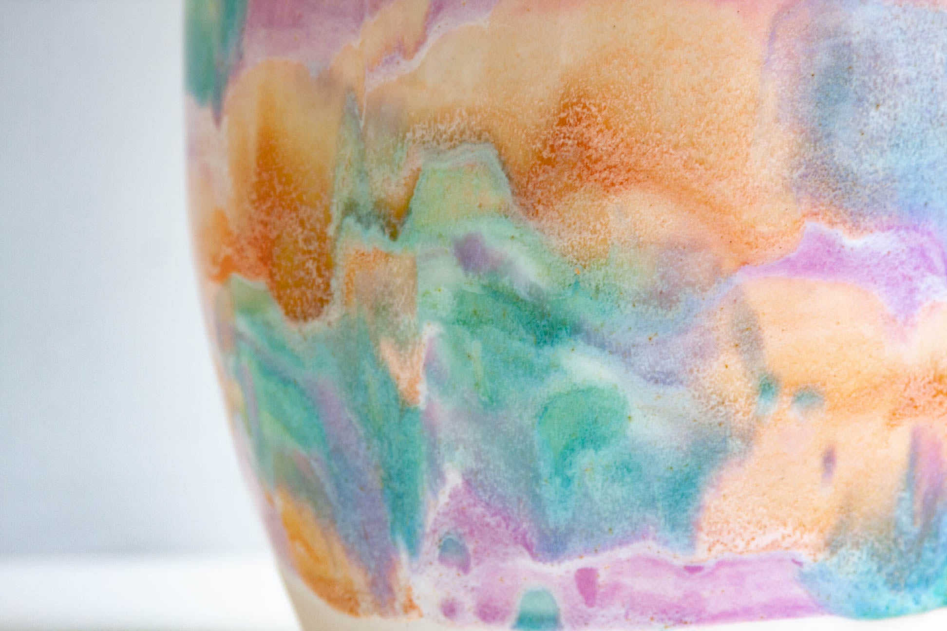 Close-up of a ceramic vase featuring a colorful watercolor wash design with blended shades of orange, teal, purple, and pink on a white base.
