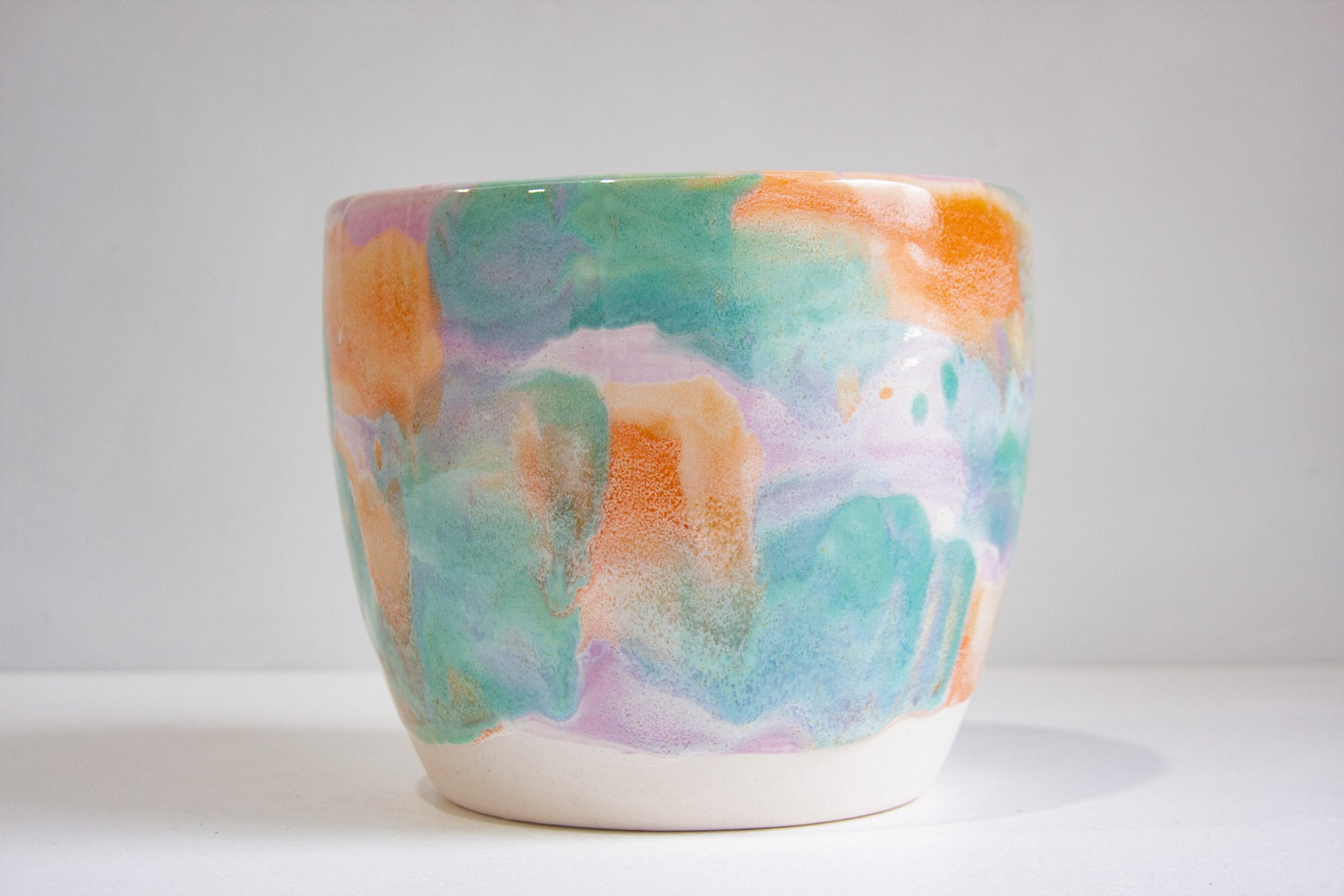A handmade ceramic planter pot with a colorful abstract watercolor glaze in shades of teal, orange, purple, and pink on a white base, set against a plain light gray background.