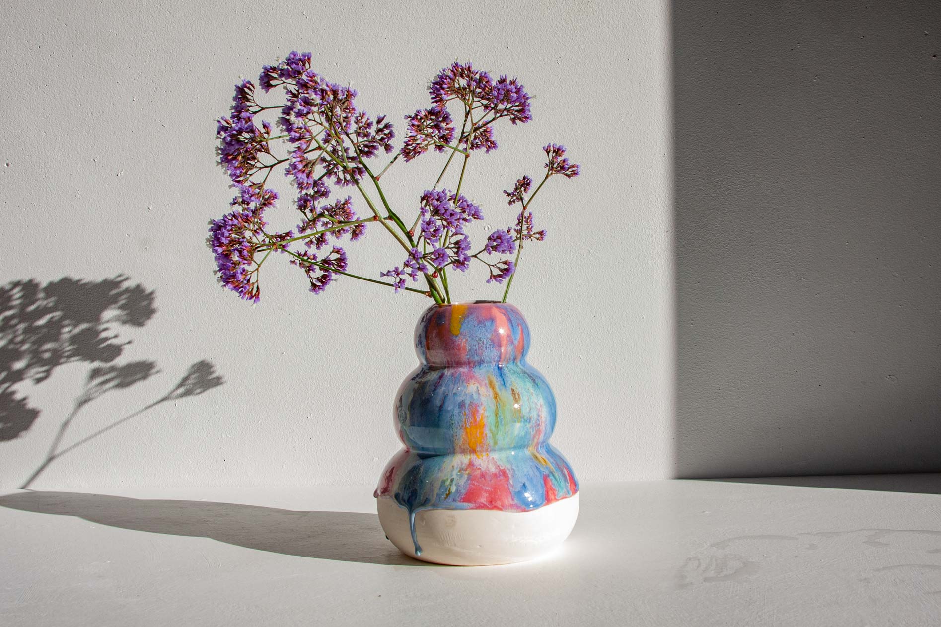 A small ceramic vase with a rounded, stacked-bottle silhouette, painted in a multicolor drip pattern of blue, pink, purple, and white. It is filled with delicate purple flowers and placed on a white surface against a white wall with a soft shadow cast to the left.