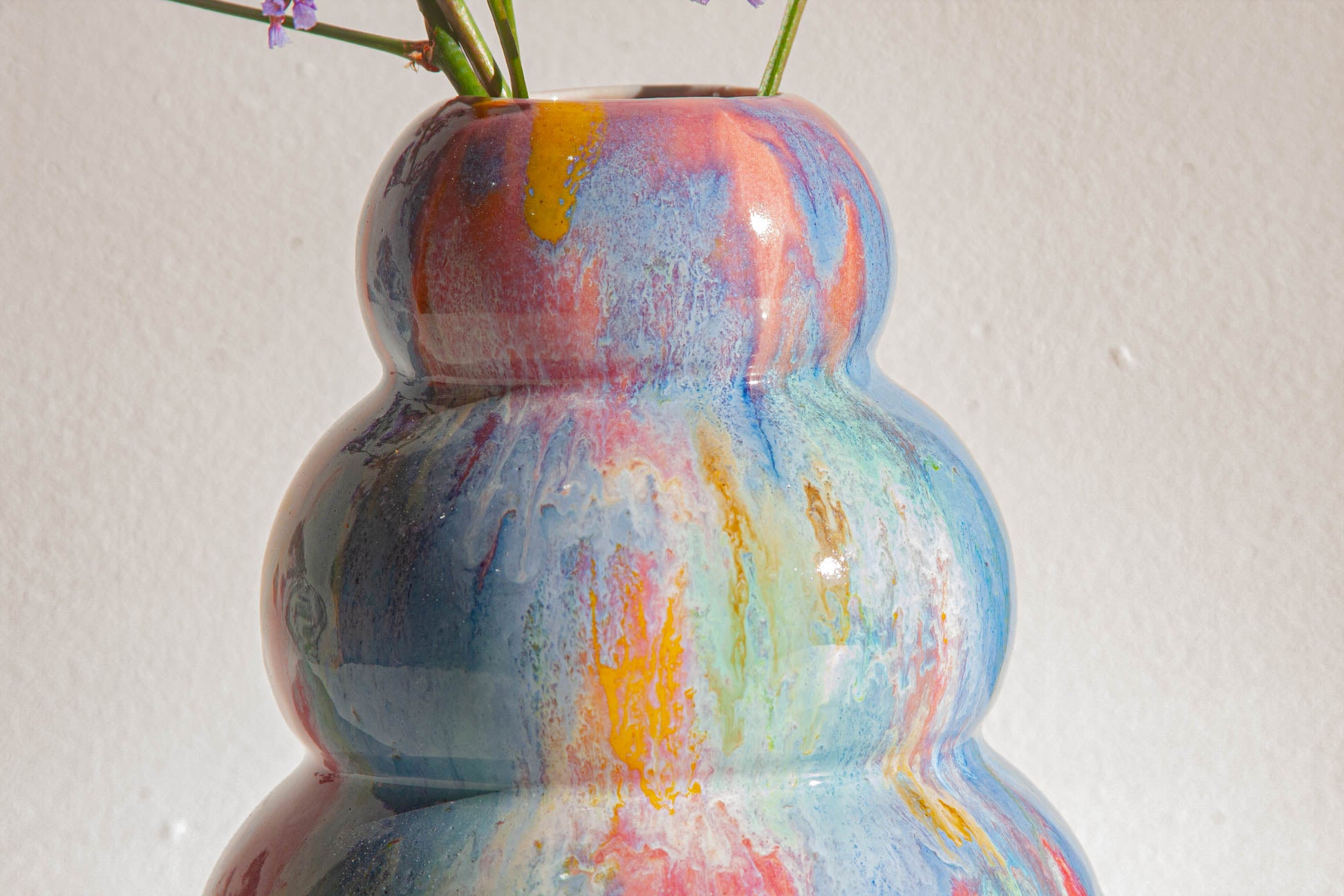 A glossy ceramic vase with a rounded, tiered shape in a marbled swirl of blue, pink, orange, and green, holding small purple flowers against a plain off-white wall.