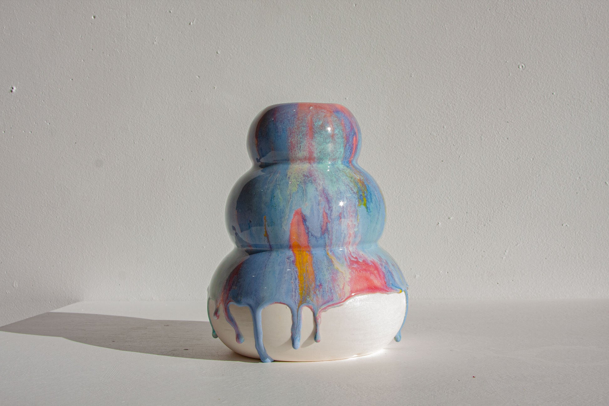 A ceramic vase with a three-tiered snowman design, featuring a glossy blue, pink, and orange drip-glaze finish on a white base, set against a plain off-white wall.