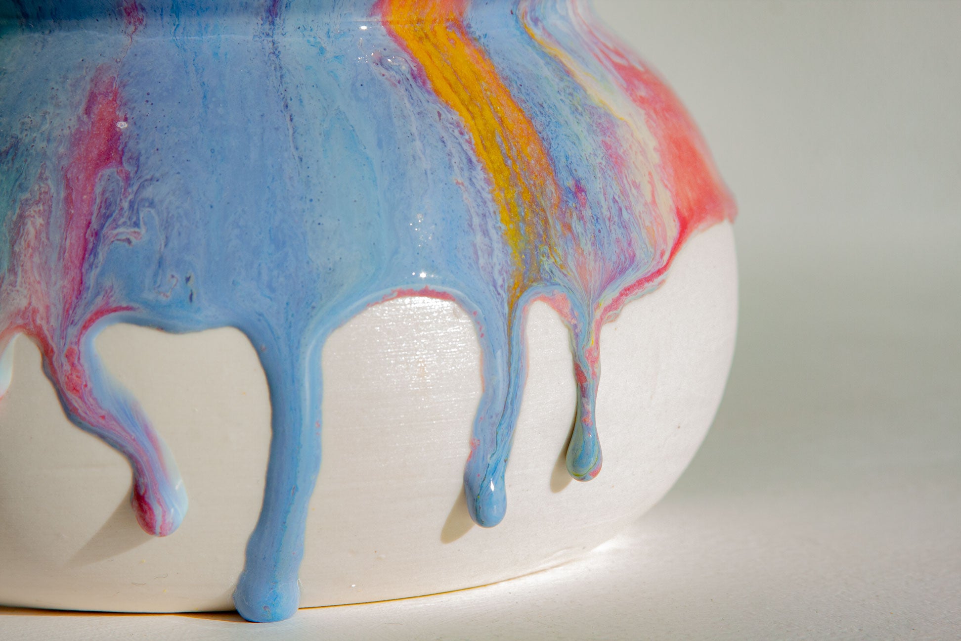 A close-up of a glossy ceramic pot with a white base and vibrant dripping paint in shades of blue, pink, purple, and yellow.